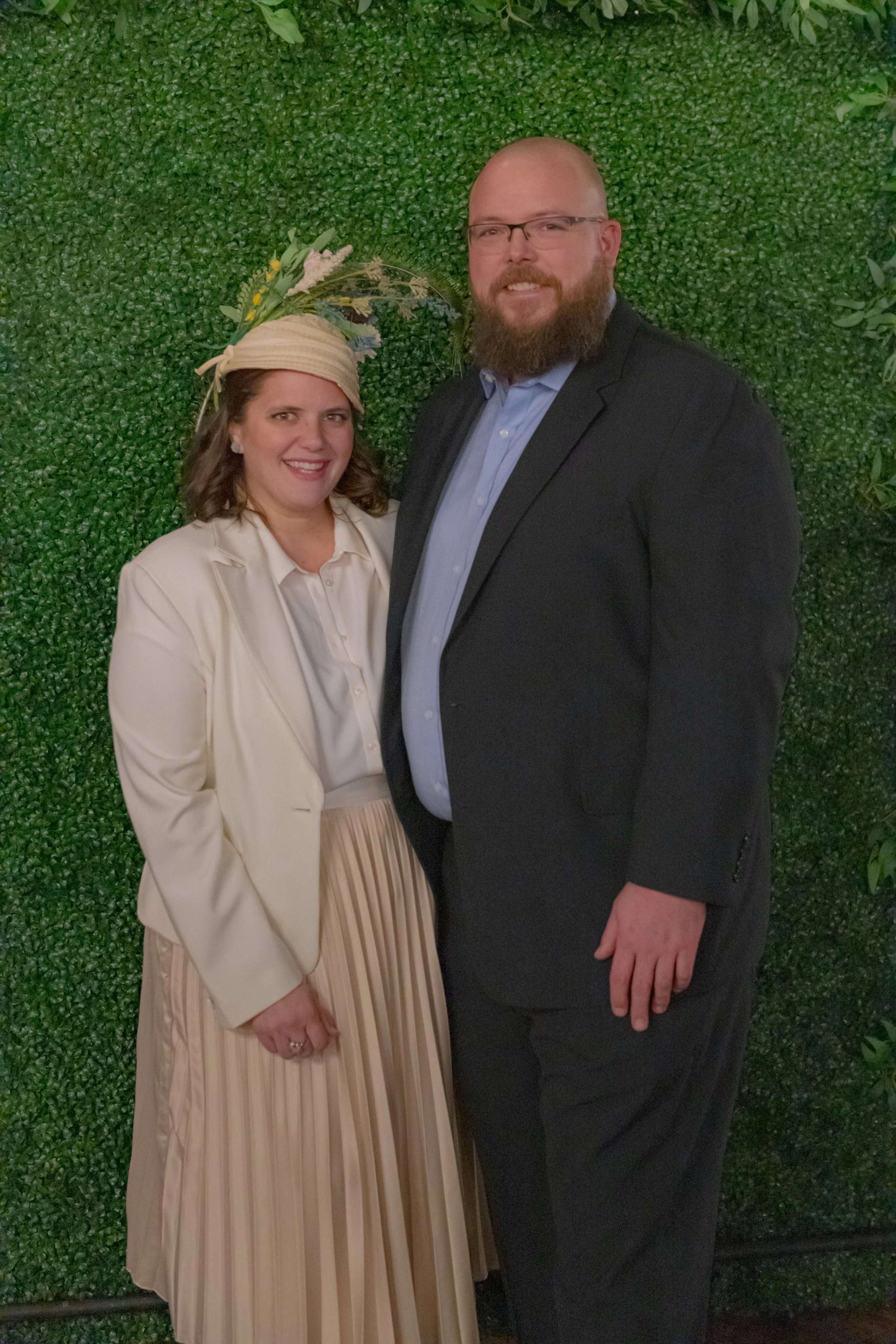 Closer portrait of two people against a green foliage backdrop, one in a cream jacket and decorative hat and one in a dark suit.