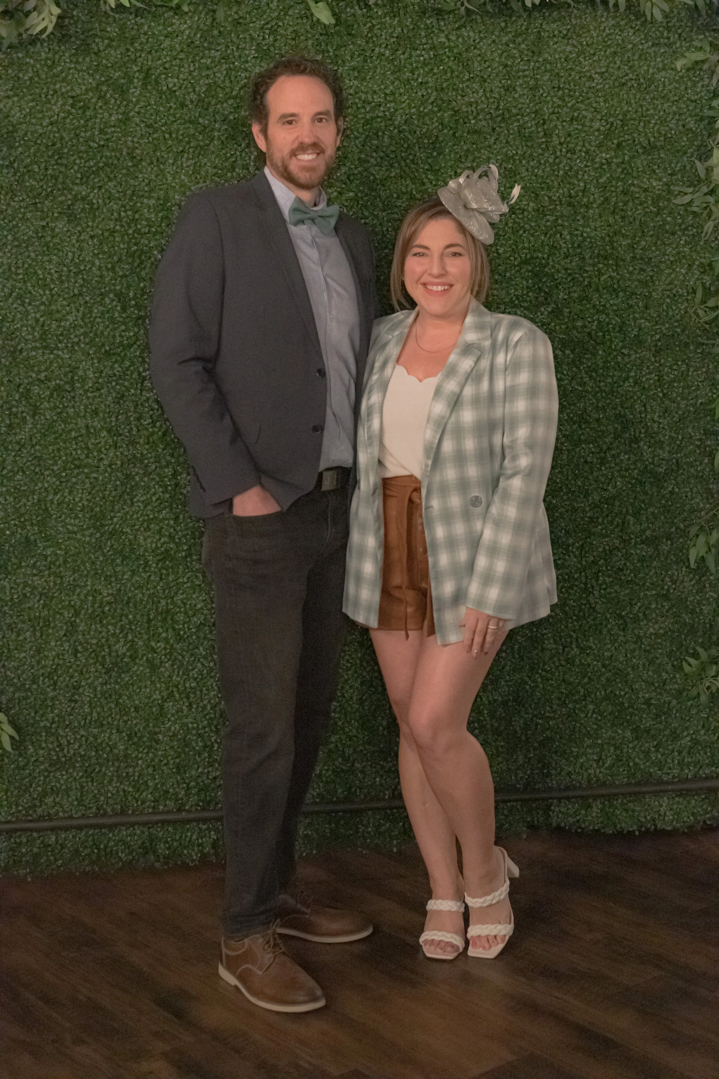 Two people pose against a green foliage backdrop, one in a dark jacket and bow tie and one in a checked jacket and decorative headpiece.