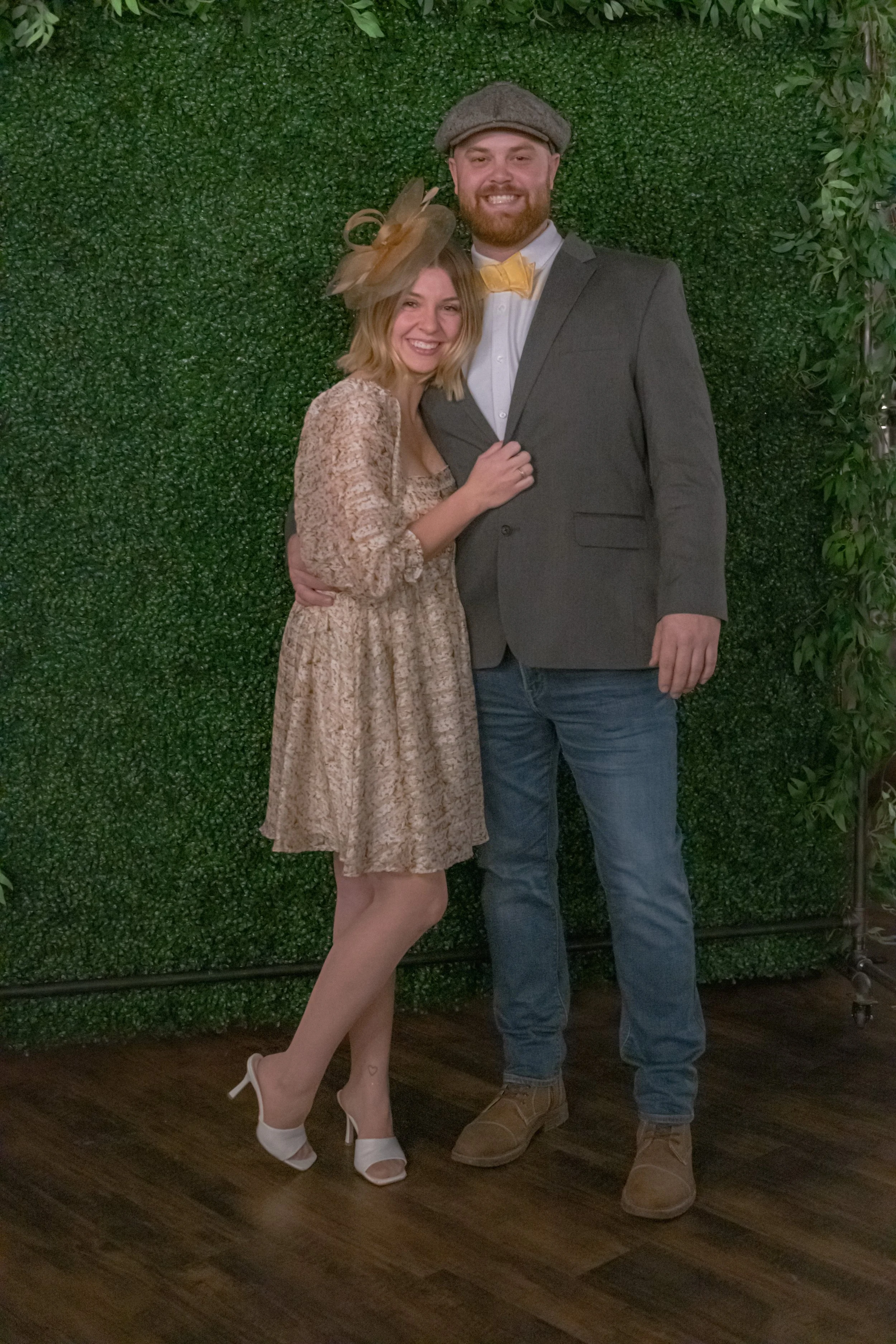 Full-length portrait of two people against a green foliage backdrop, one in a patterned dress and one in a gray jacket, flat cap and yellow bow tie.