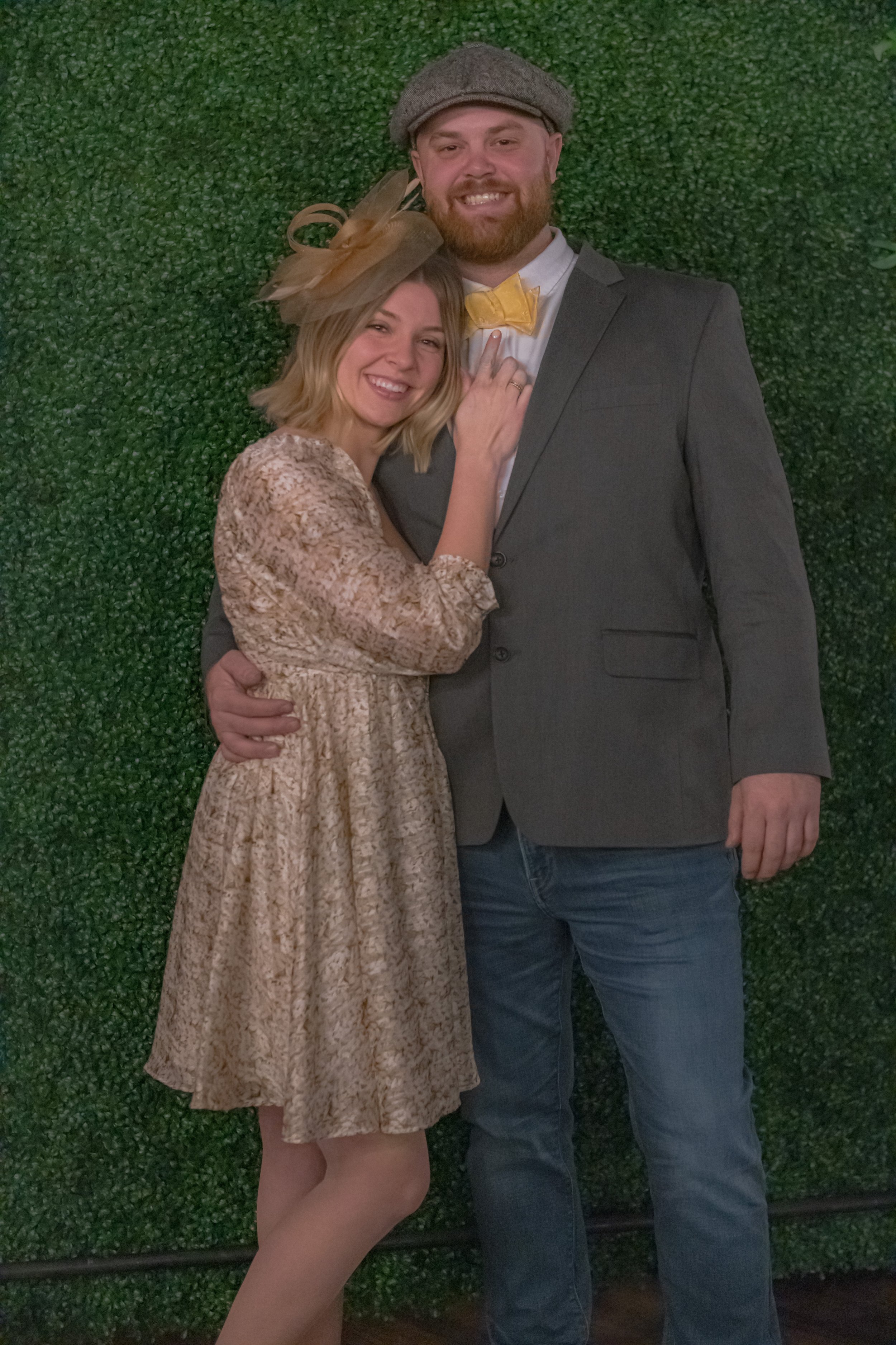 Closer portrait of two people against a green foliage backdrop, one in a patterned dress and one in a gray jacket, flat cap and yellow bow tie.