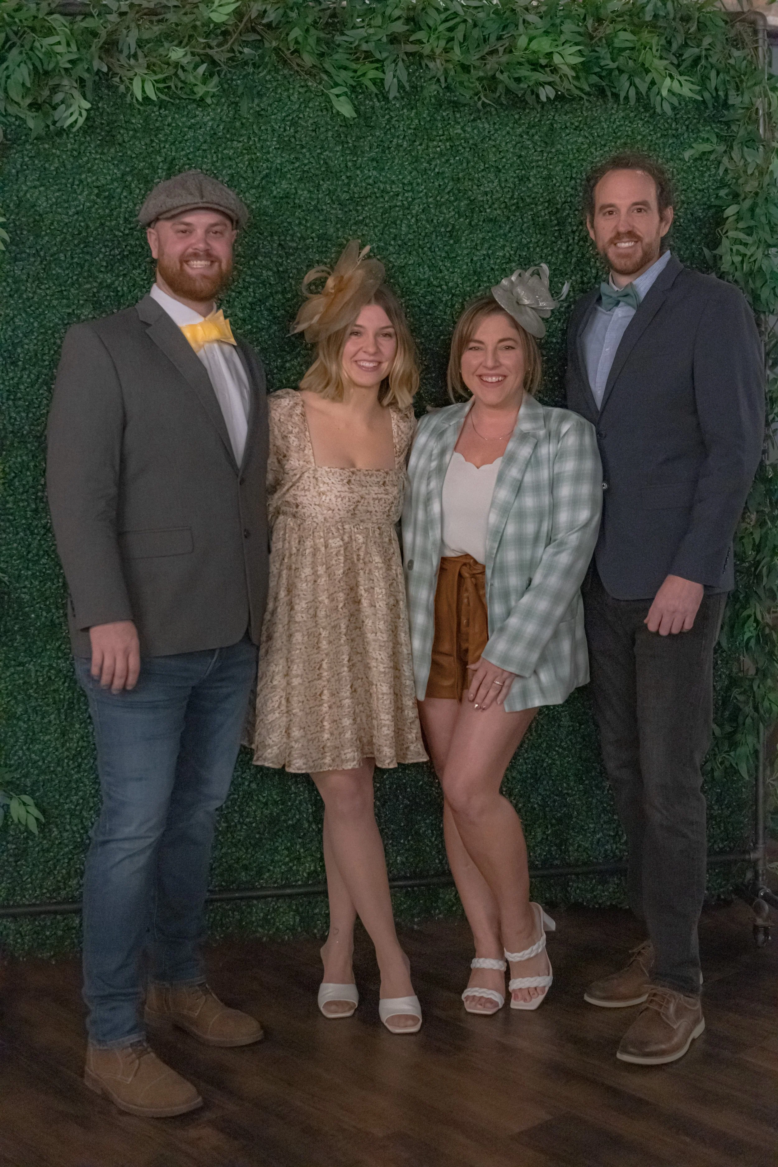 Full-length group portrait of four people in dress clothes against a green foliage backdrop.