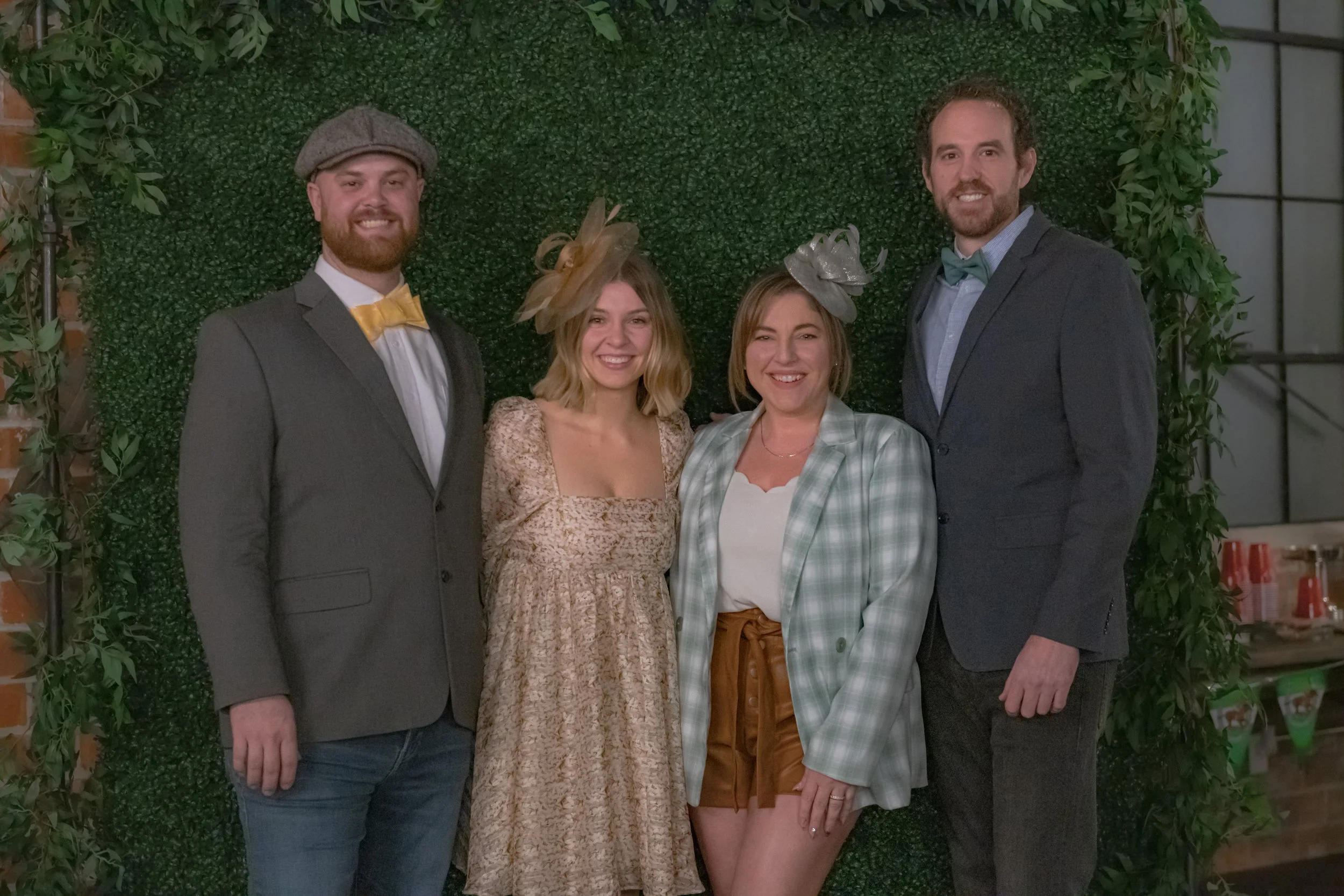 Four people pose shoulder to shoulder against a green foliage backdrop, with bow ties and decorative headpieces.