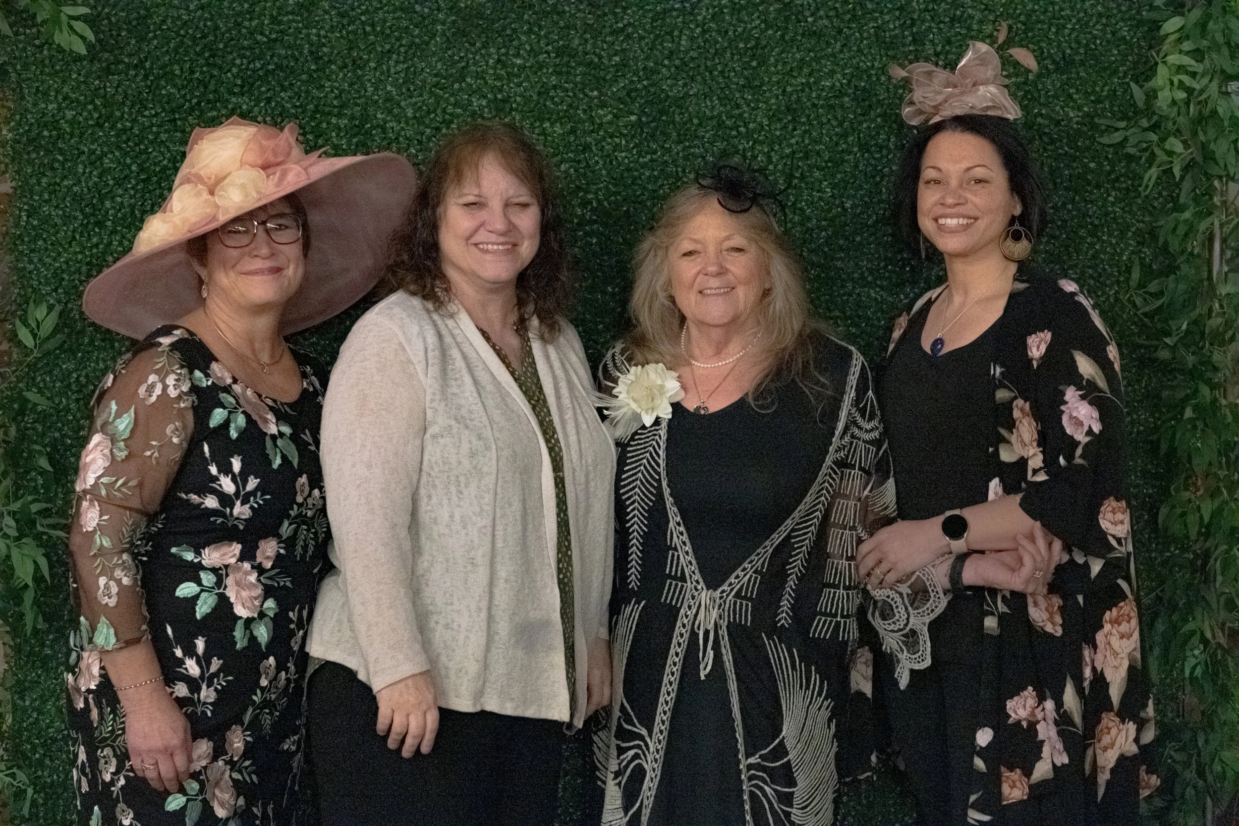 Four people in patterned outfits pose together against a green foliage backdrop, with a wide-brimmed hat at left.