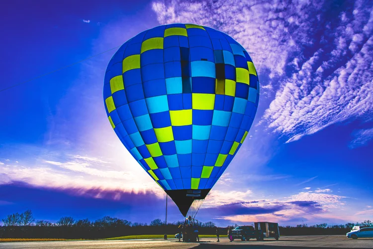 A blue and yellow-green hot-air balloon stands inflated beneath a colorful evening sky.