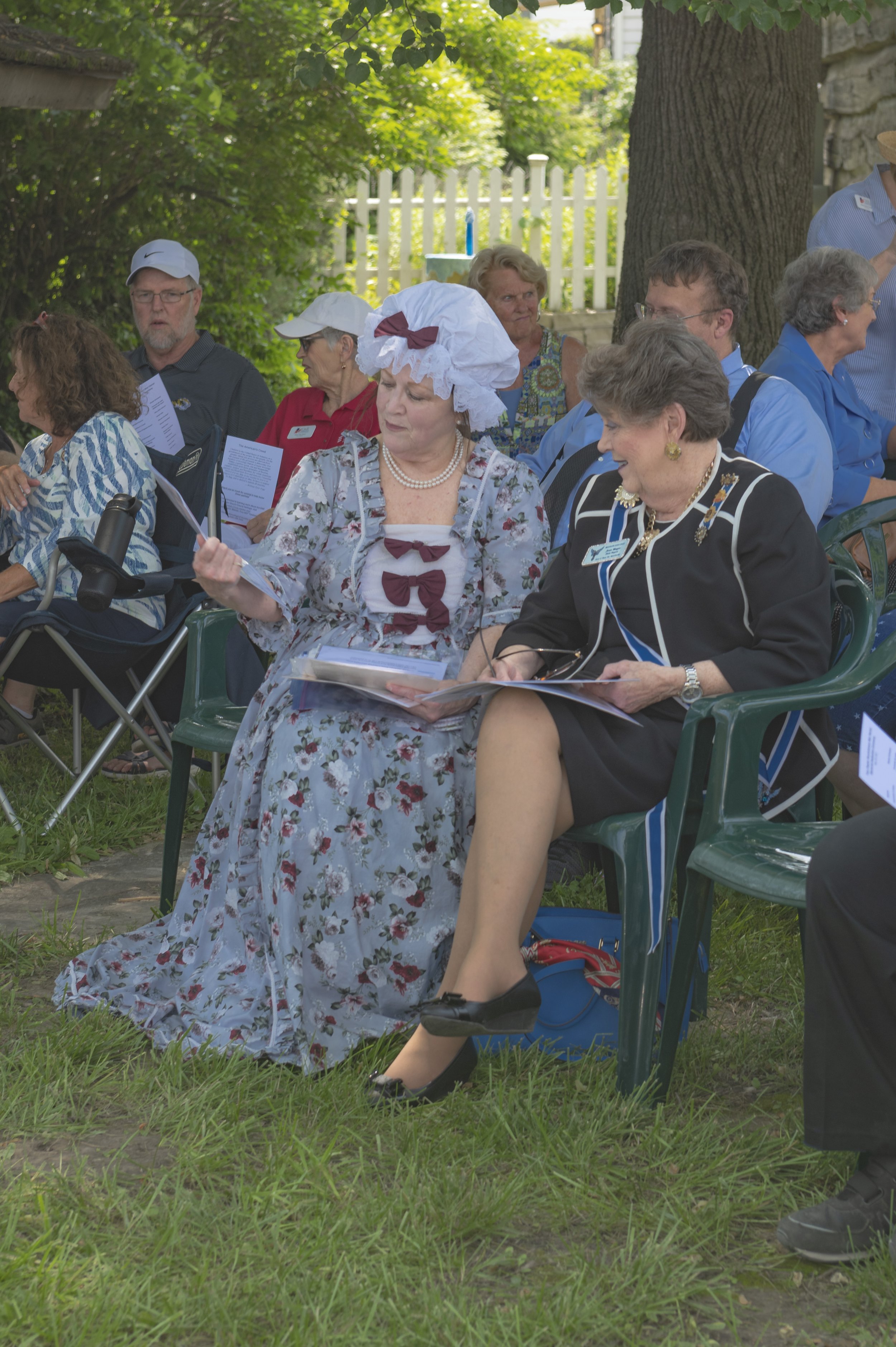 Two attendees read programs, one wearing a floral period dress and bonnet at the 2022 Daughters of the American Revolution ceremony.