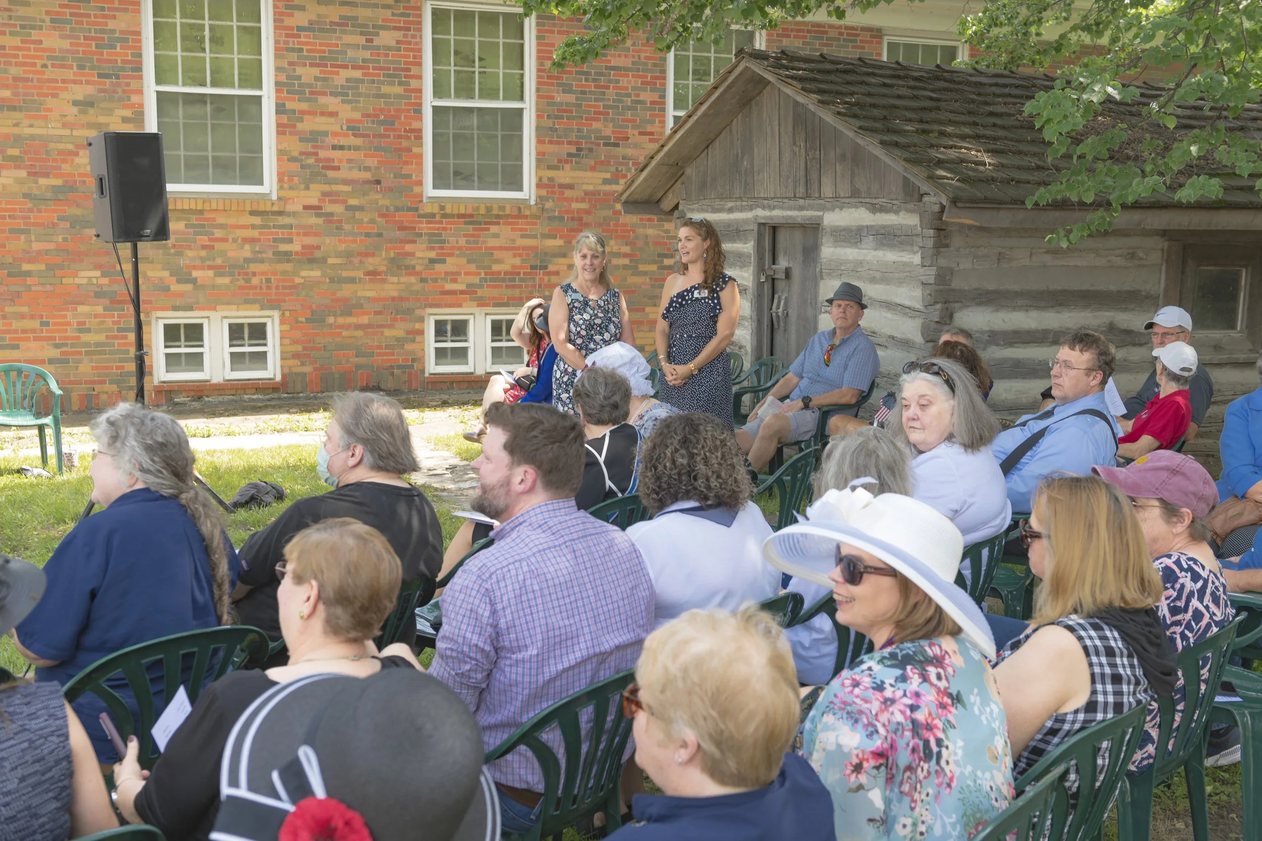 Seated guests look toward the speakers beside the brick building and log structure at the 2022 Daughters of the American Revolution ceremony.