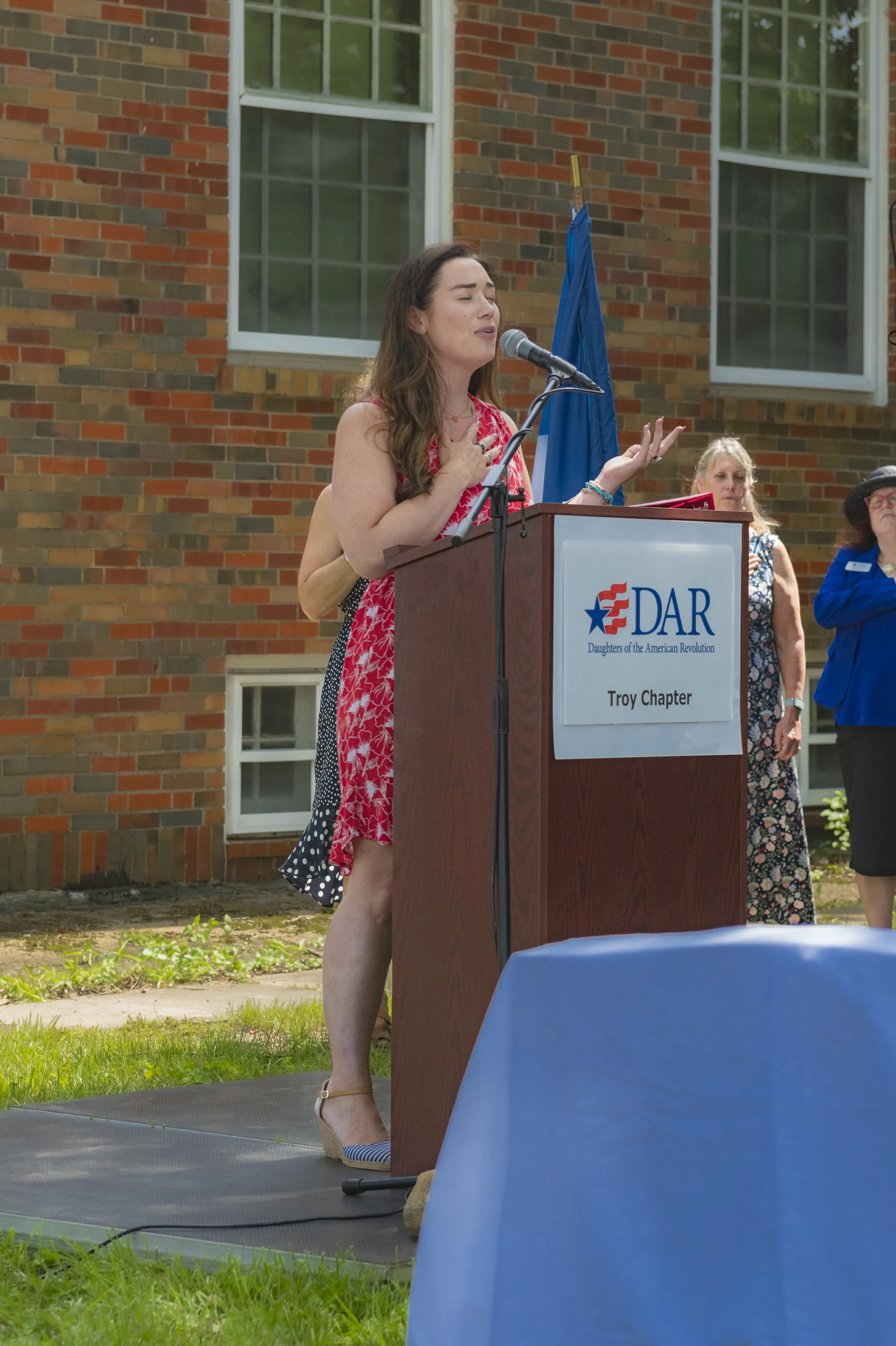 A speaker in a red floral dress addresses guests at the DAR Troy Chapter lectern at the 2022 Daughters of the American Revolution ceremony.
