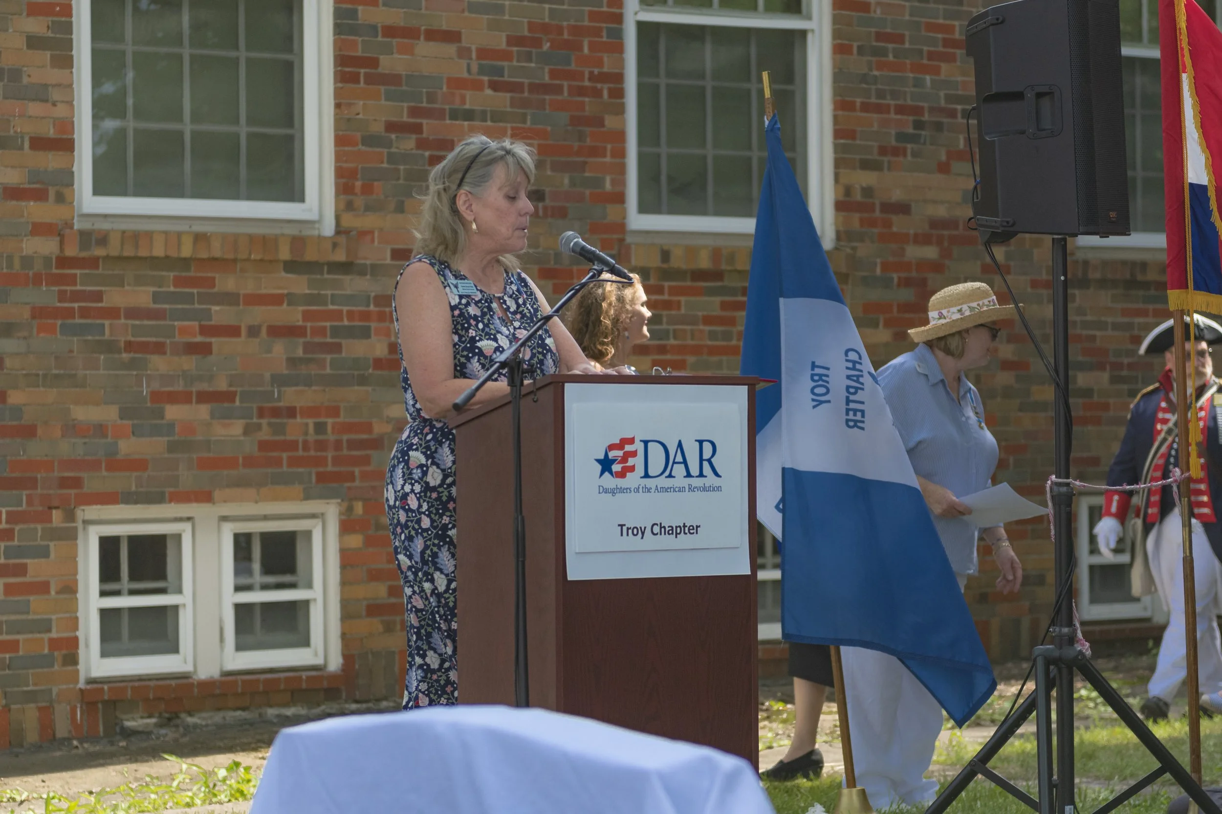 A speaker in a floral dress stands at the lectern beside the chapter flag at the 2022 Daughters of the American Revolution ceremony.