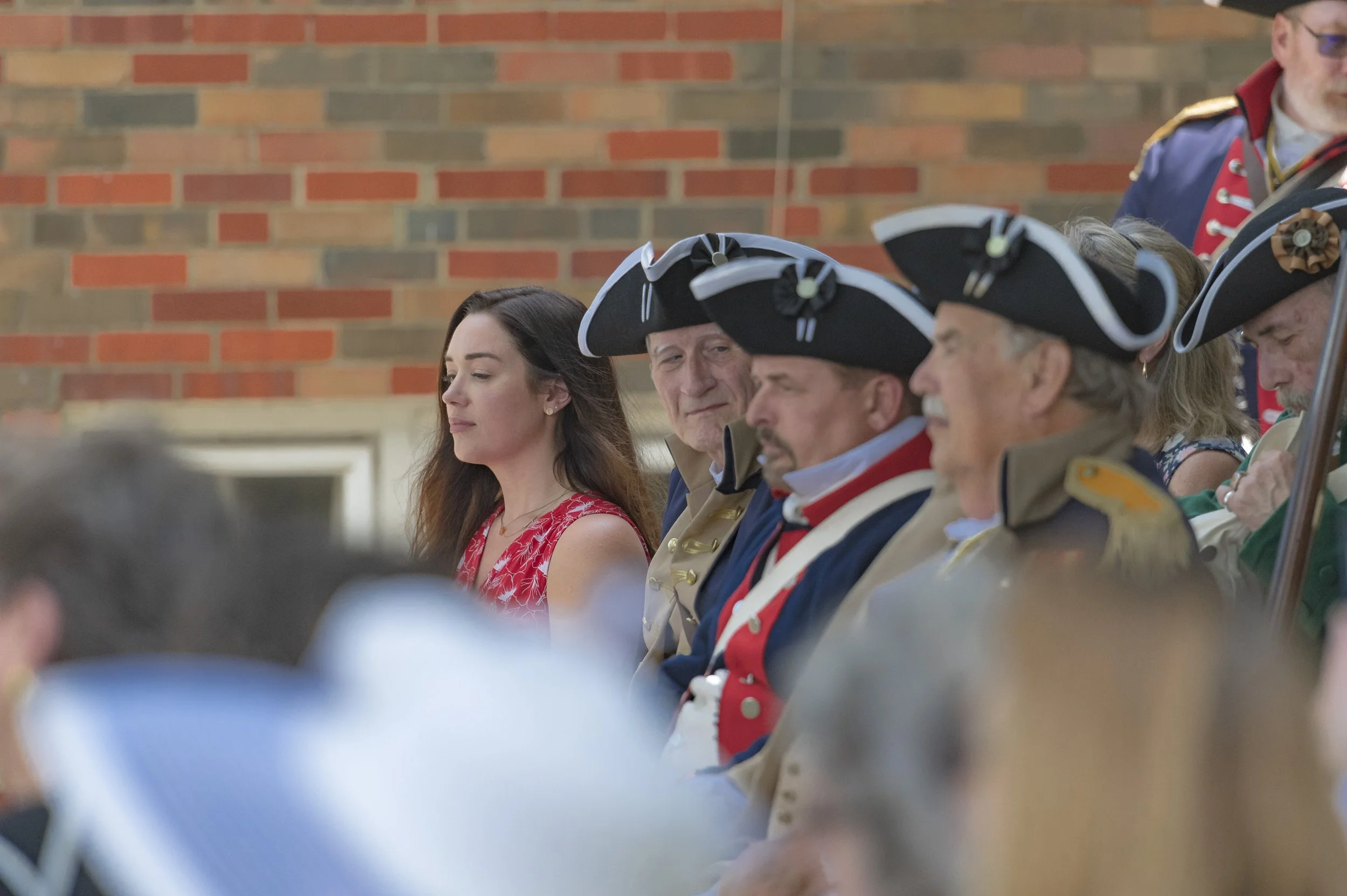 Participants in period uniforms sit beside a guest in a red floral dress at the 2022 Daughters of the American Revolution ceremony.
