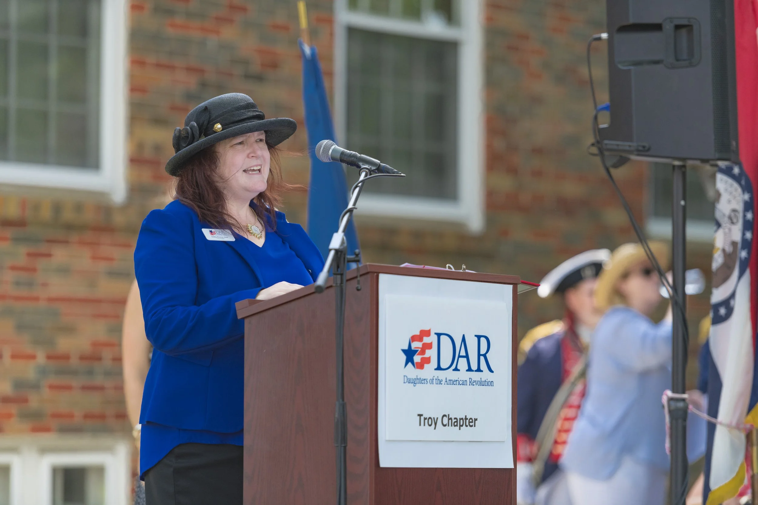 A speaker in a blue jacket and black hat addresses the audience at the 2022 Daughters of the American Revolution ceremony.