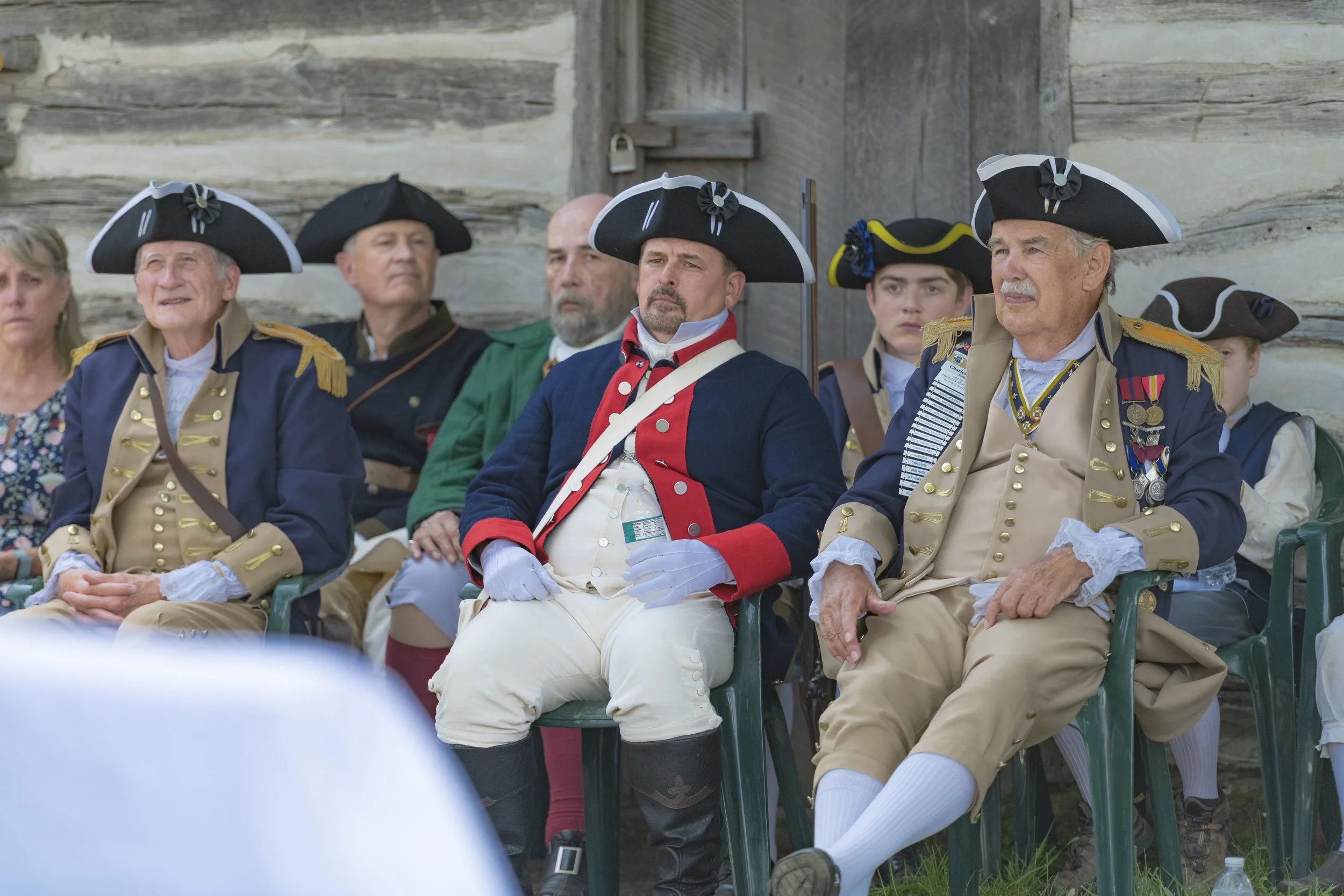 Participants in period clothing sit in a row beside the log structure at the 2022 Daughters of the American Revolution ceremony.