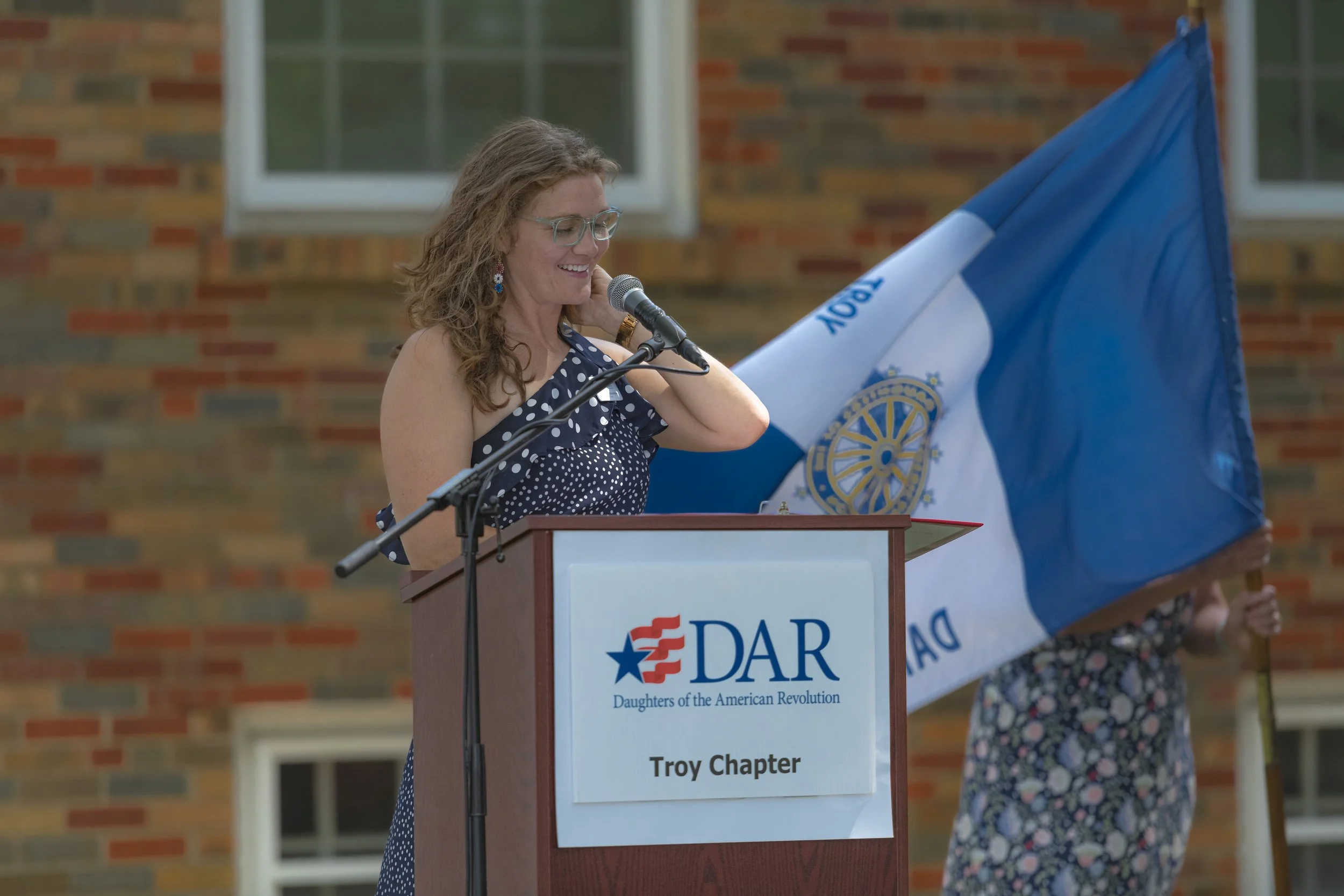 A speaker in a navy patterned dress stands beside the blue-and-white chapter flag at the 2022 Daughters of the American Revolution ceremony.