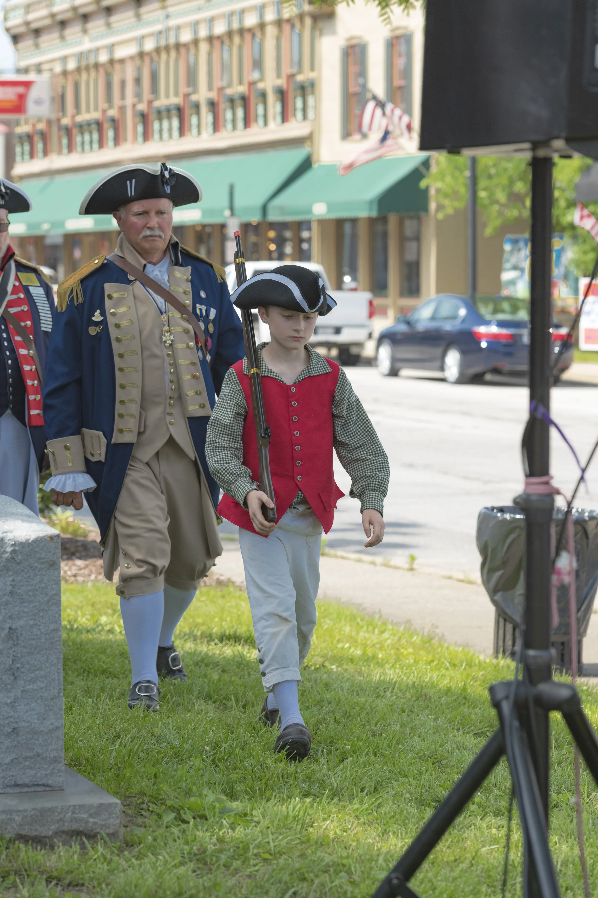 A young participant in a red waistcoat walks ahead of an adult in period clothing at the 2022 Daughters of the American Revolution ceremony.