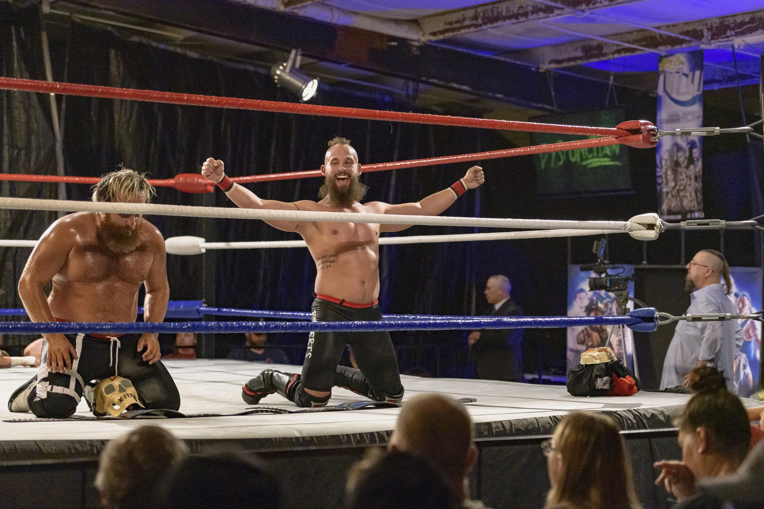 Two wrestlers kneel on the mat beside a gold belt, one raising both arms at the 2022 World League Wrestling event.