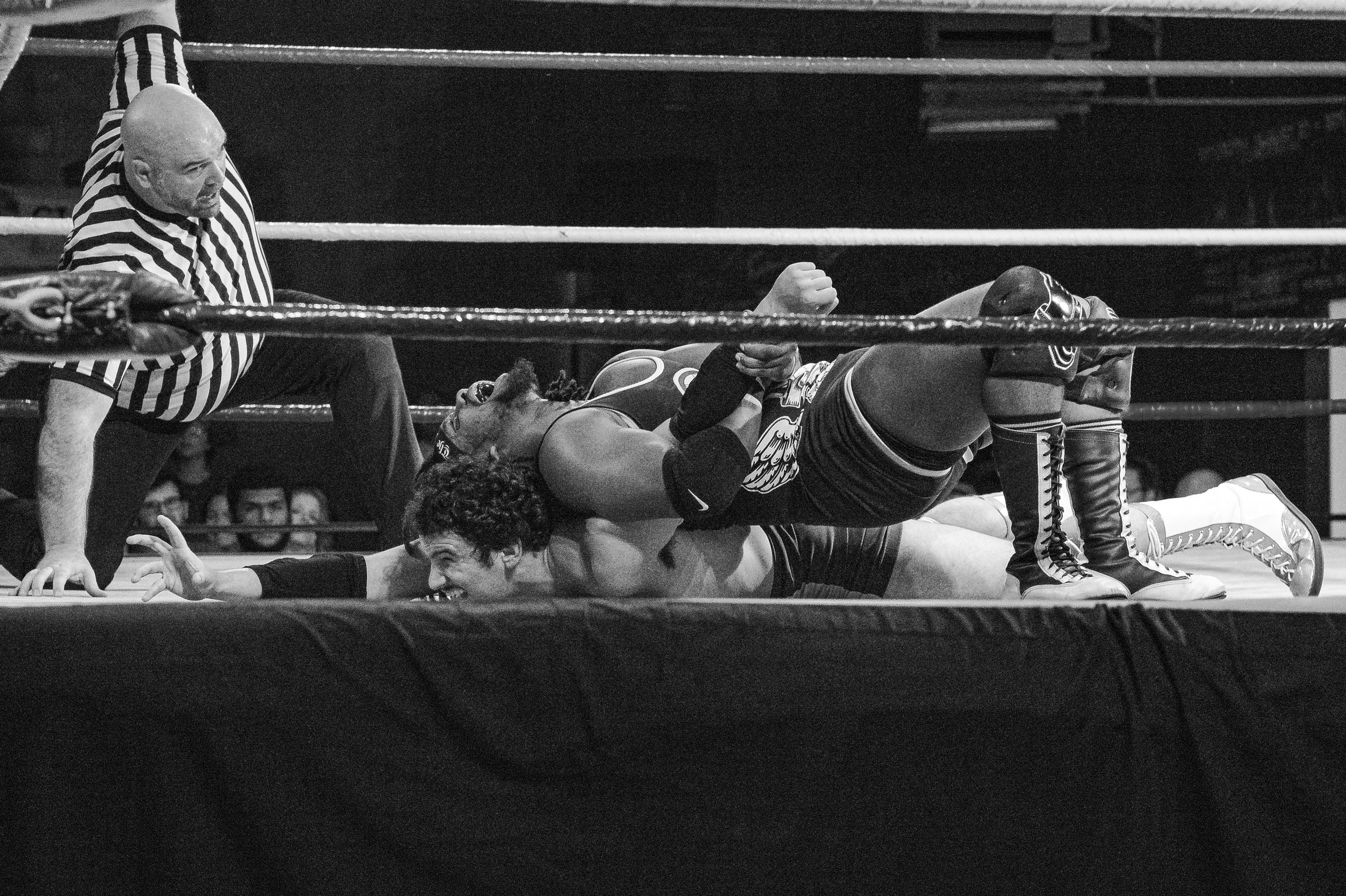 A black-and-white photograph shows a referee watching closely as two wrestlers grapple on the mat at the 2022 World League Wrestling event.