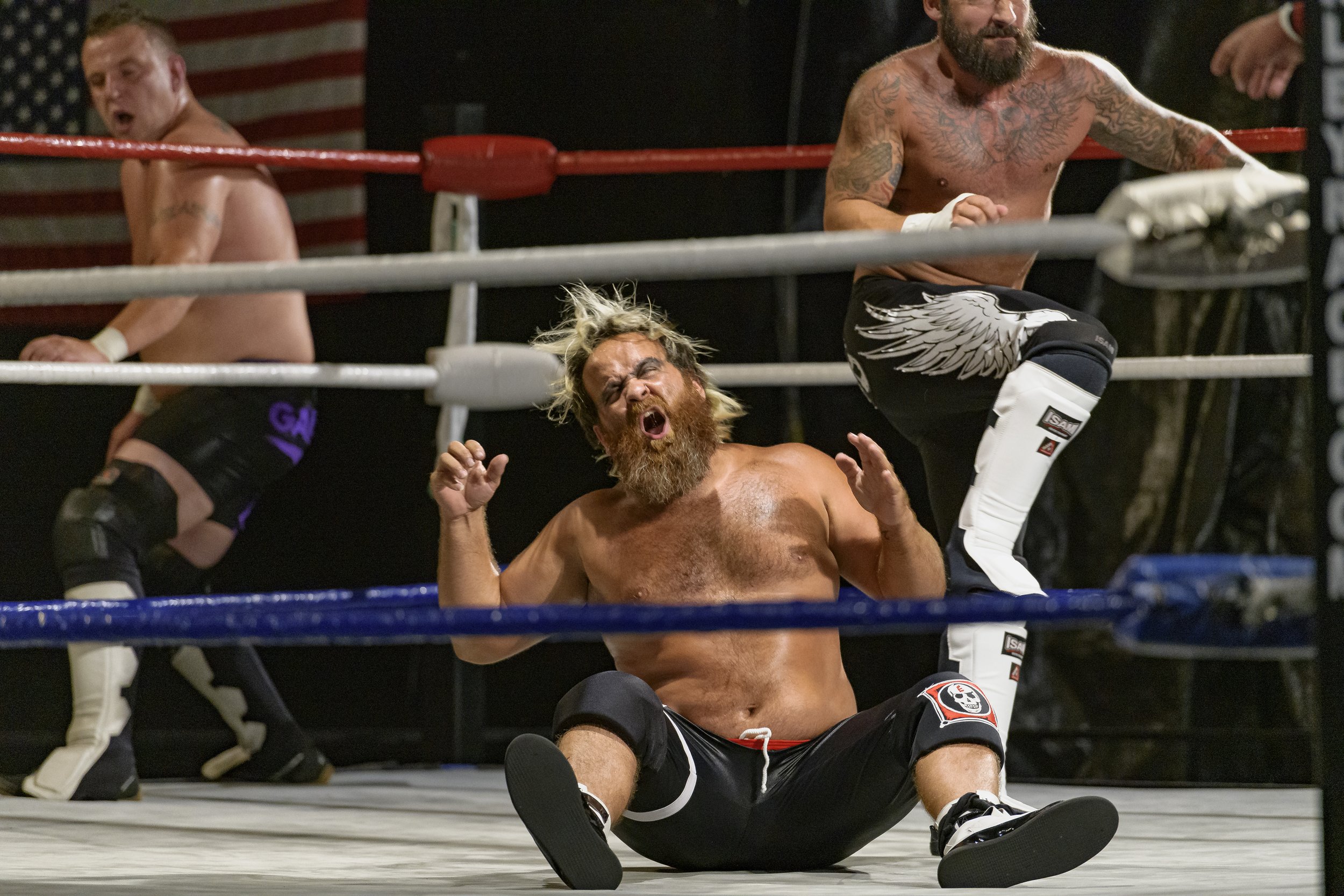 A wrestler sits on the mat with another standing behind him near the ropes at the 2022 World League Wrestling event.