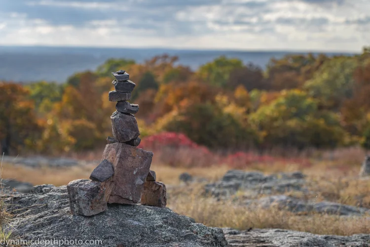 A small stack of stones stands on a rocky ledge above autumn-colored trees.