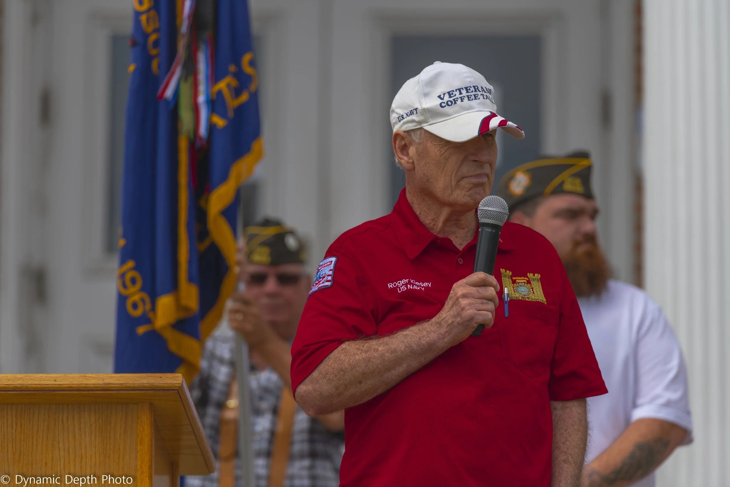 A speaker wearing a red shirt and white cap holds a microphone during the 2021 Memorial Day Service.