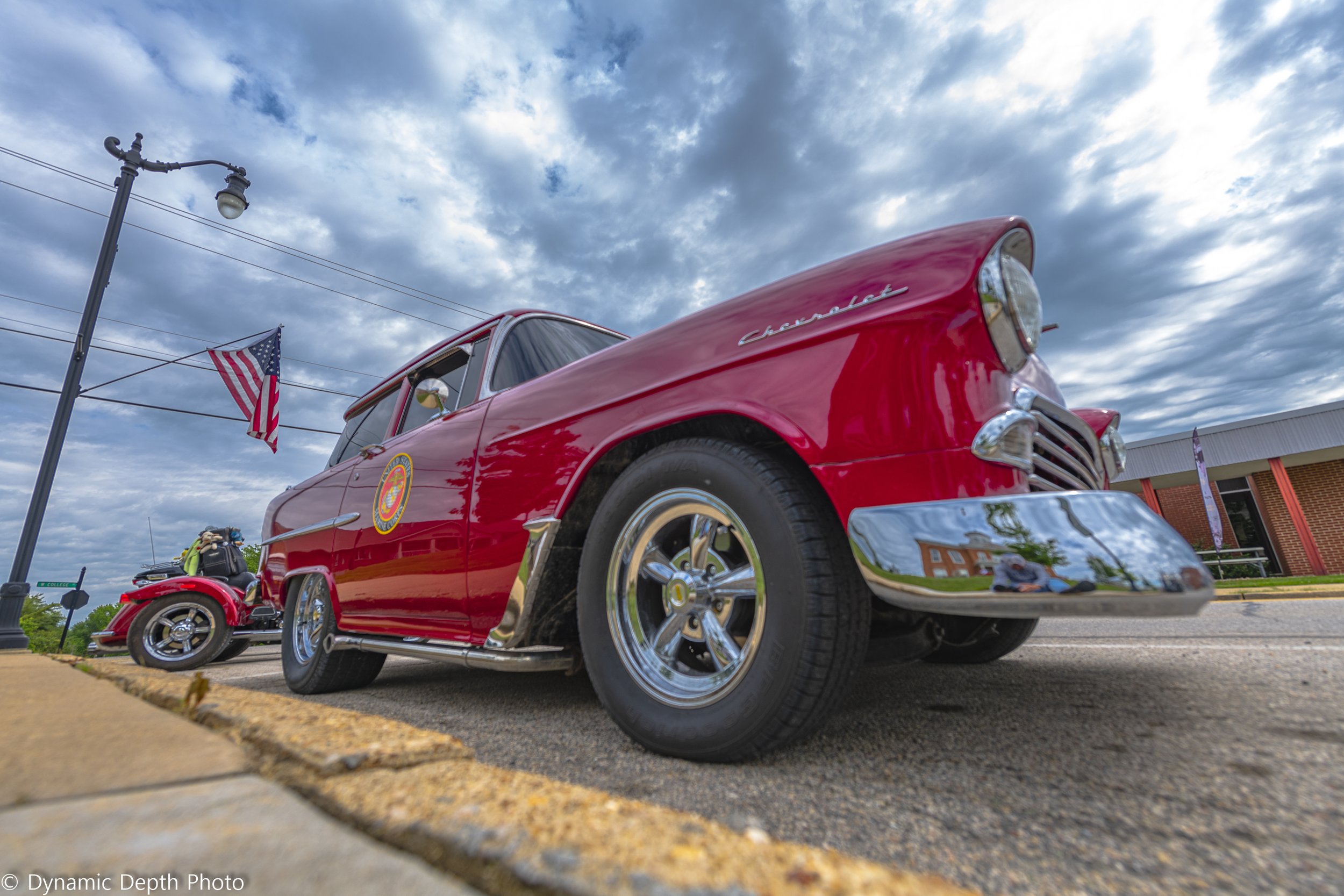 A low-angle view shows a red vintage Chevrolet and an American flag beneath a cloudy sky.