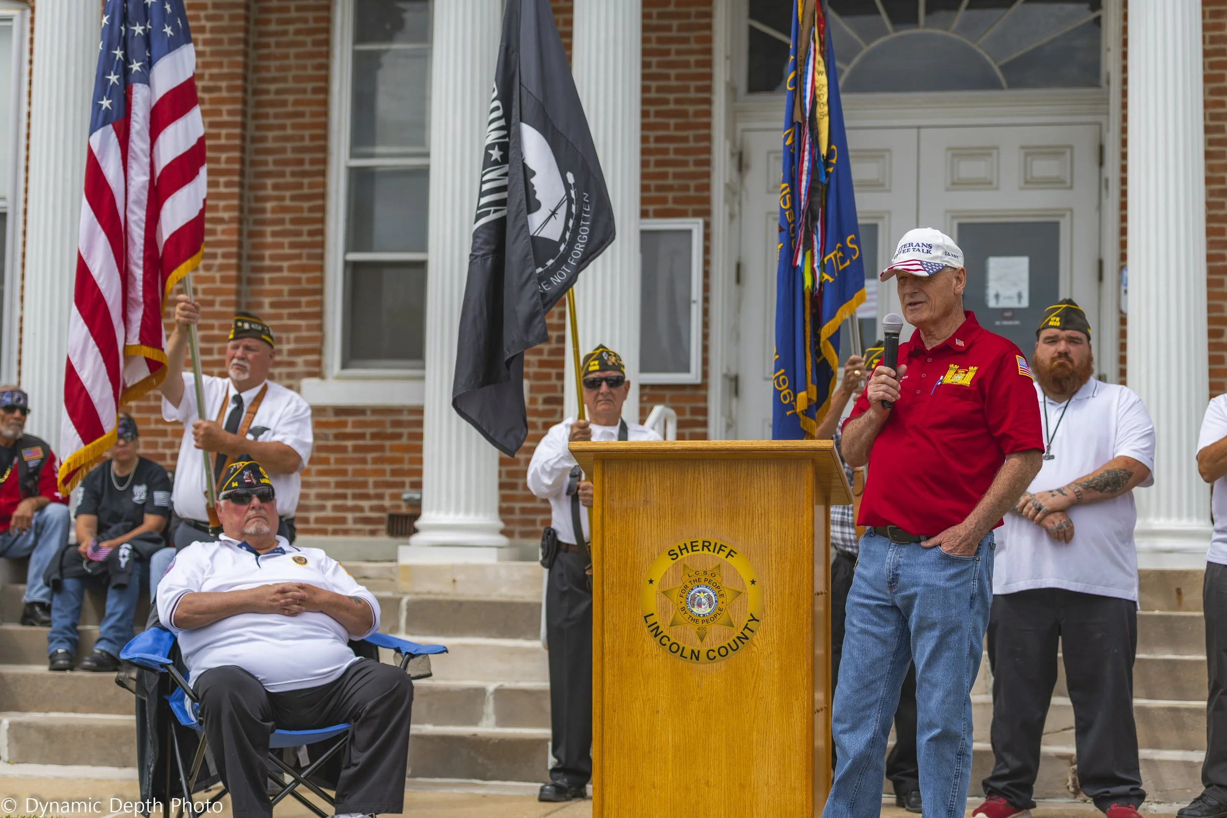 A speaker in a red shirt addresses the gathering while participants sit and stand beside the lectern.