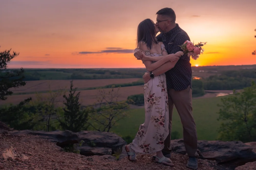 A couple embraces above a wide valley beneath an orange sunset.