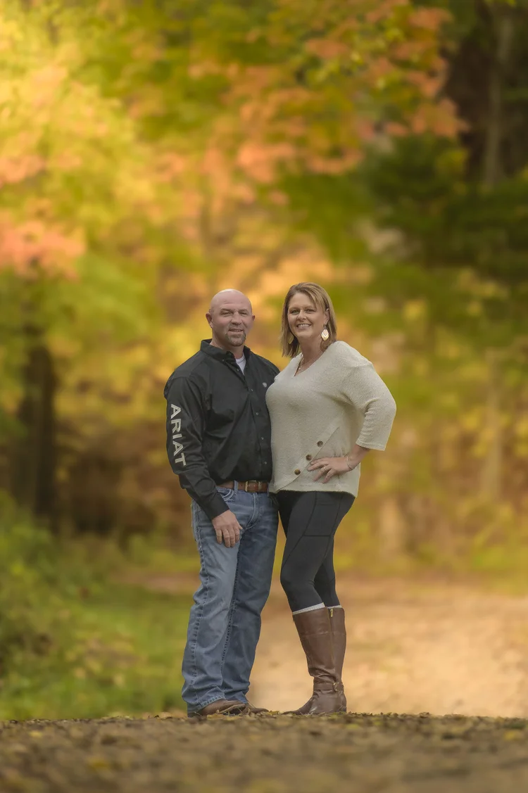 A couple stands on a woodland path surrounded by yellow and green autumn leaves.