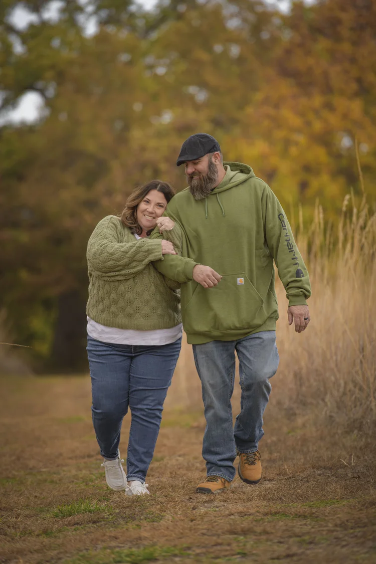A couple walks together along a path bordered by tall grass and autumn foliage.