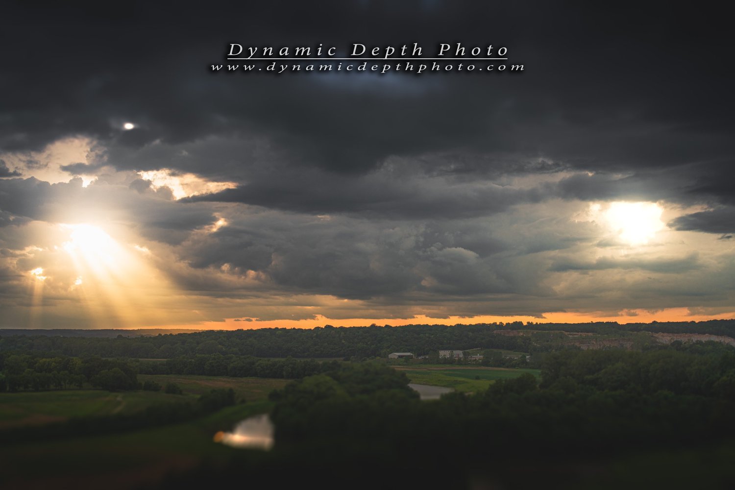 Sun rays breaking through dark, heavy clouds over a rural landscape with trees, fields, and a small body of water.