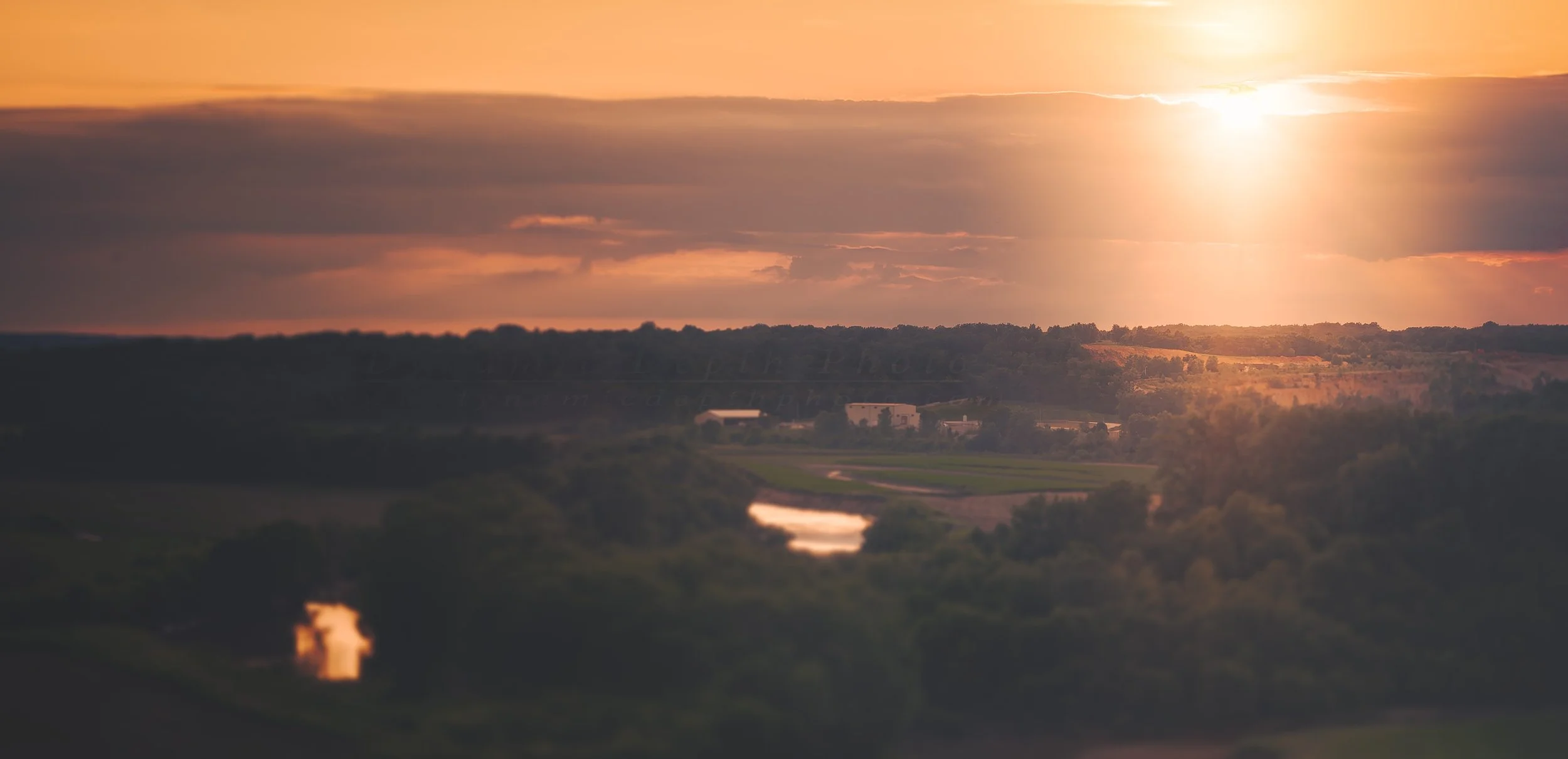 Warm evening light breaks through a bank of clouds, spreading across wooded hills and open fields. Below, the winding water catches the orange glow while the foreground settles into shadow.