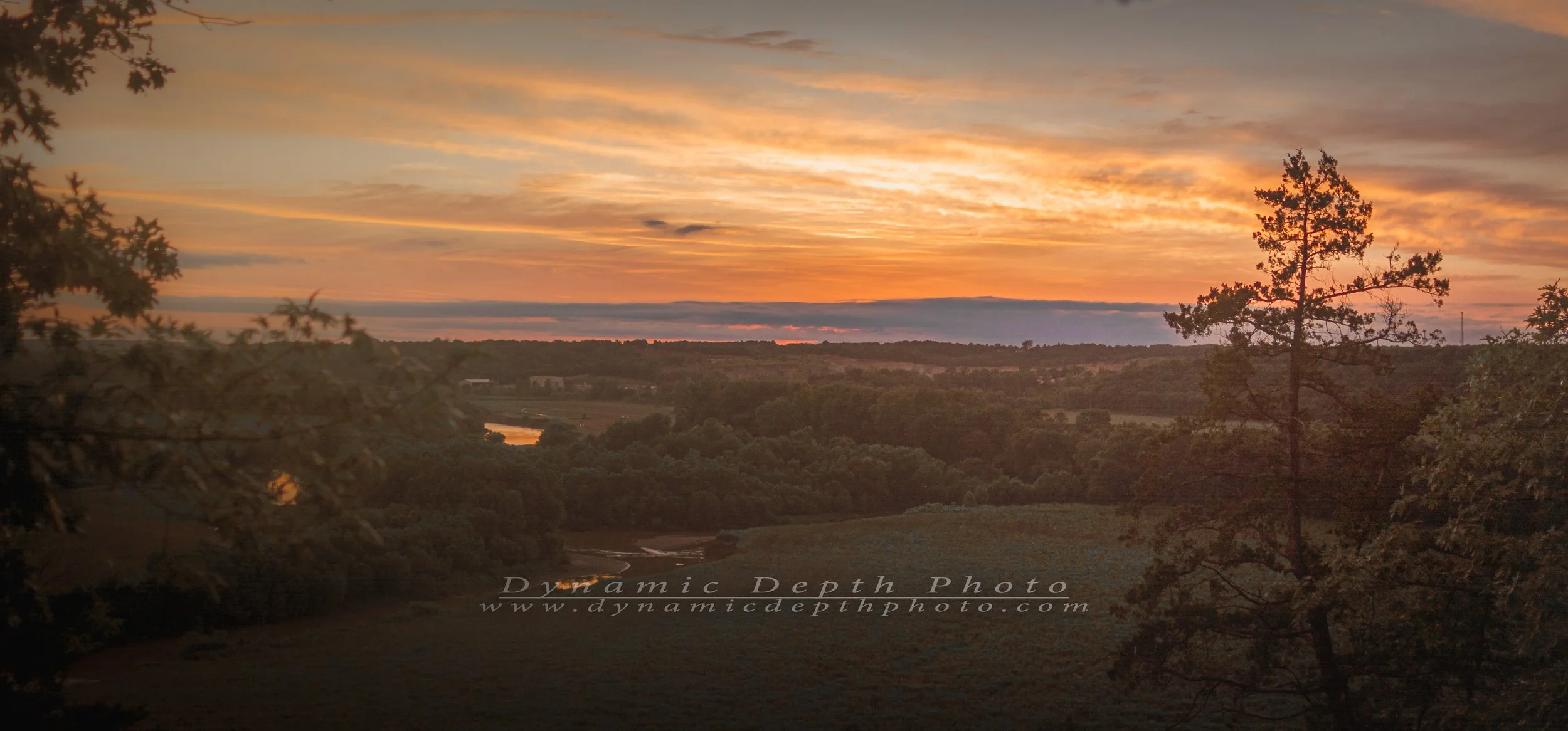 Orange and gold clouds stretch across the evening sky above wooded hills and open fields. Framed by trees, the overlook reveals small reflections of sunset on the water below as the landscape settles into shadow.