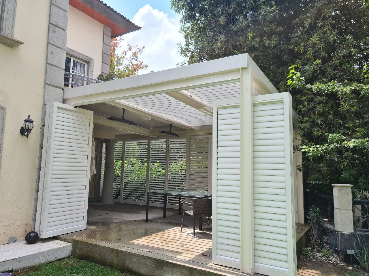 Outdoor pergola with white louvered walls and ceiling attached to a house and overlooking a garden