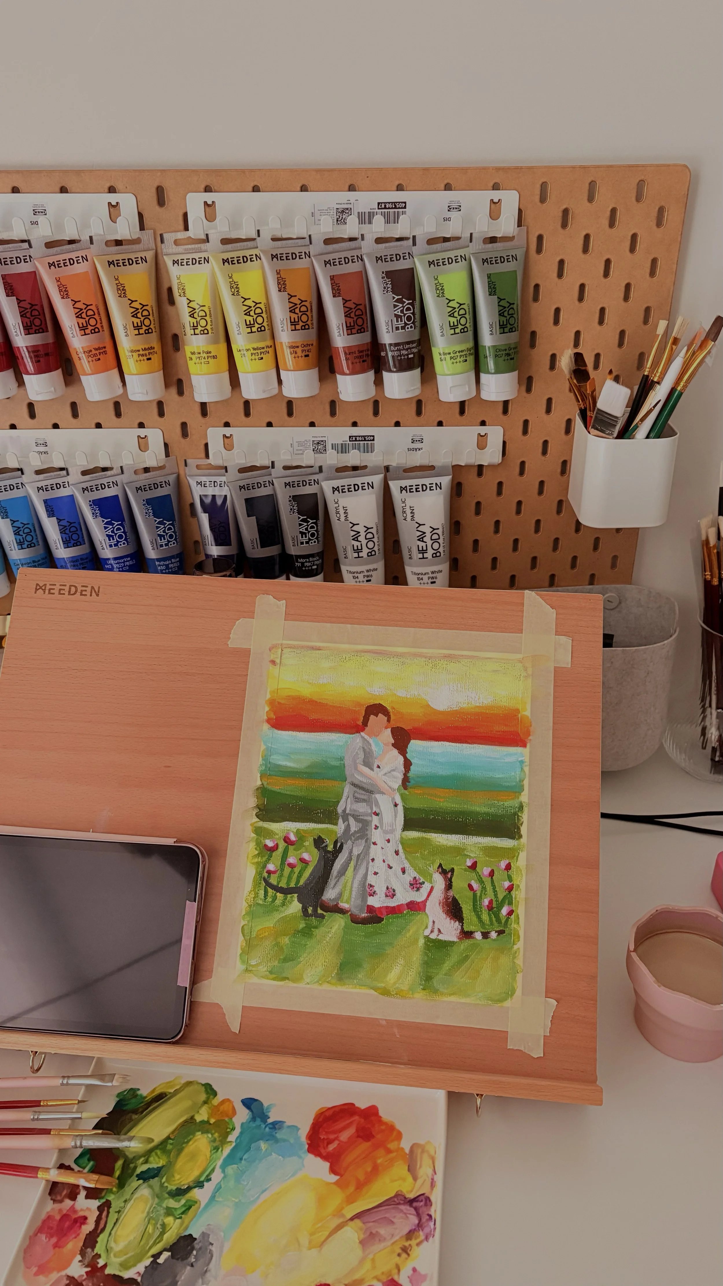 A painting of a couple standing together on a grassy field with a sunset sky in the background, two cats near them, and a garden with flowers in the foreground, taped to a wooden art board.