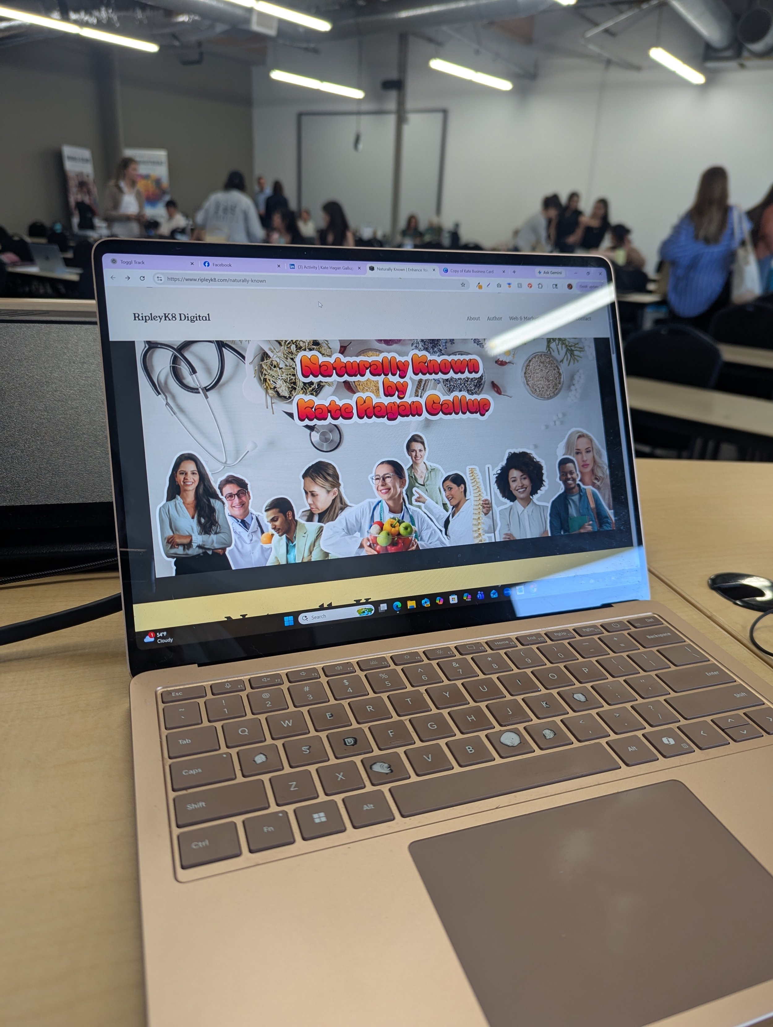 A laptop computer on a table in a classroom or conference room filled with students and people.