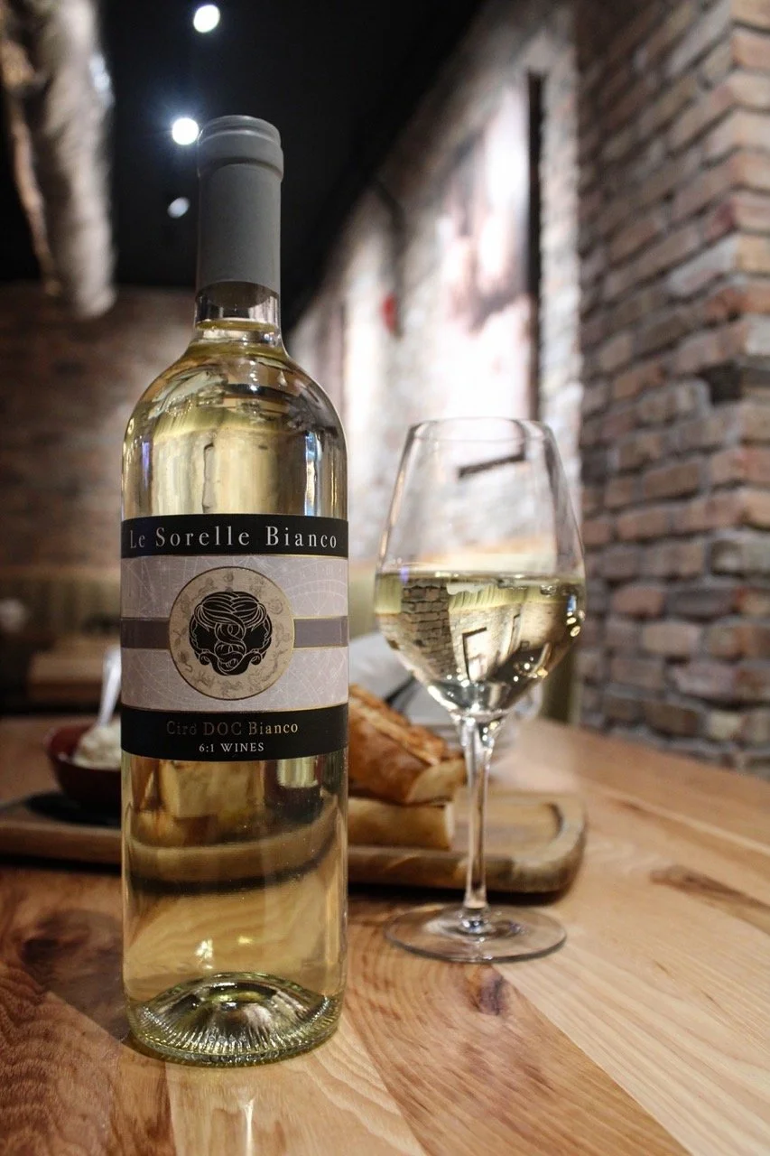 A bottle of white wine with a glass of wine beside it on a wooden table in a cozy, rustic setting with brick walls.