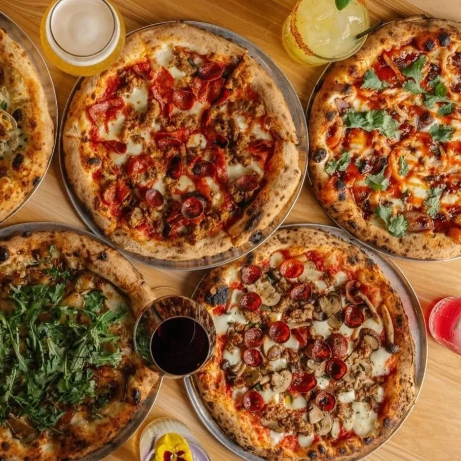 Four pizzas with various toppings on a wooden table, along with drinks including a glass of beer, a glass of wine, and a soda, and a yellow beverage with a lime wedge.