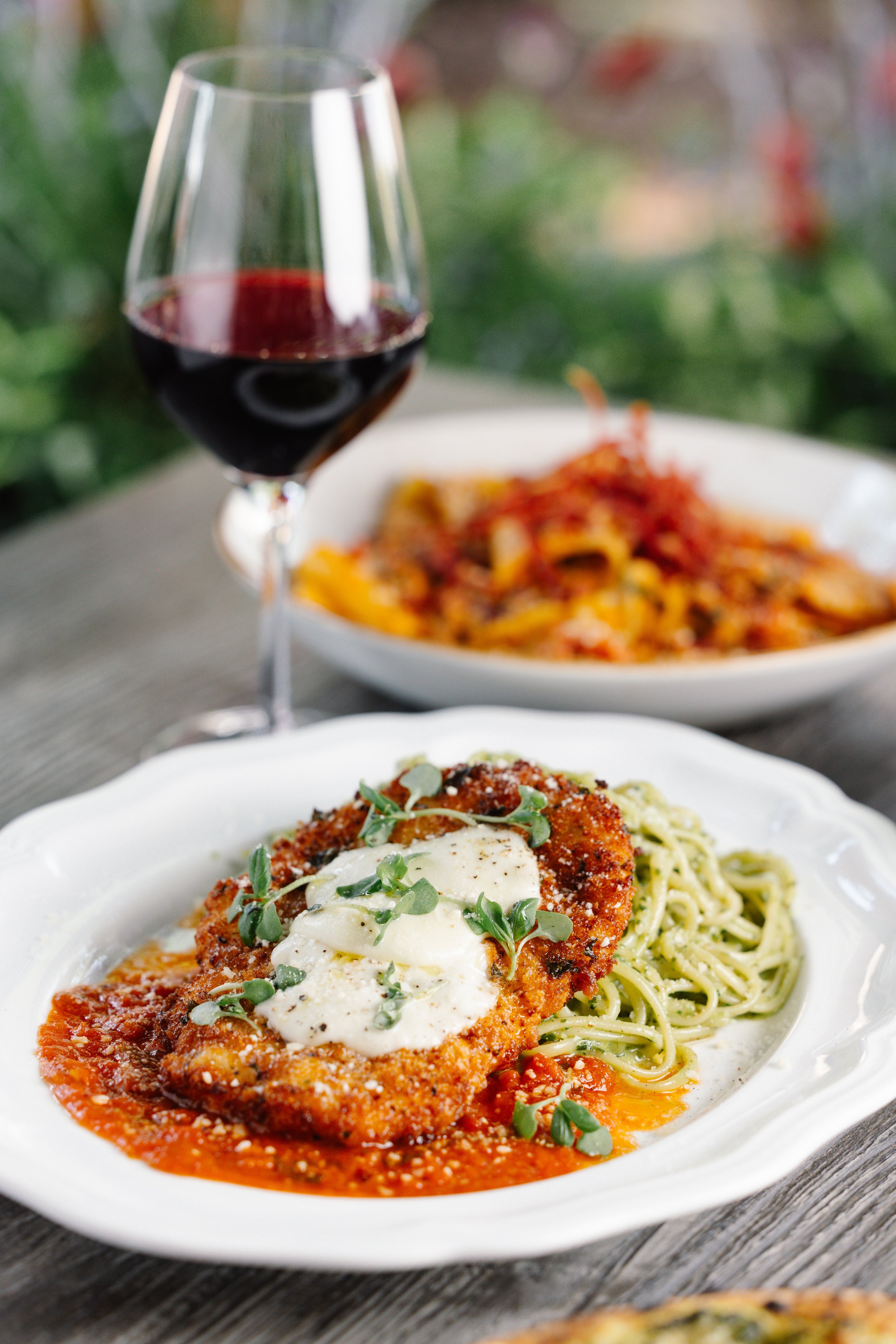 Plate of breaded chicken topped with sauce, cheese, and herbs beside a serving of spaghetti, with a glass of red wine and another dish in the background