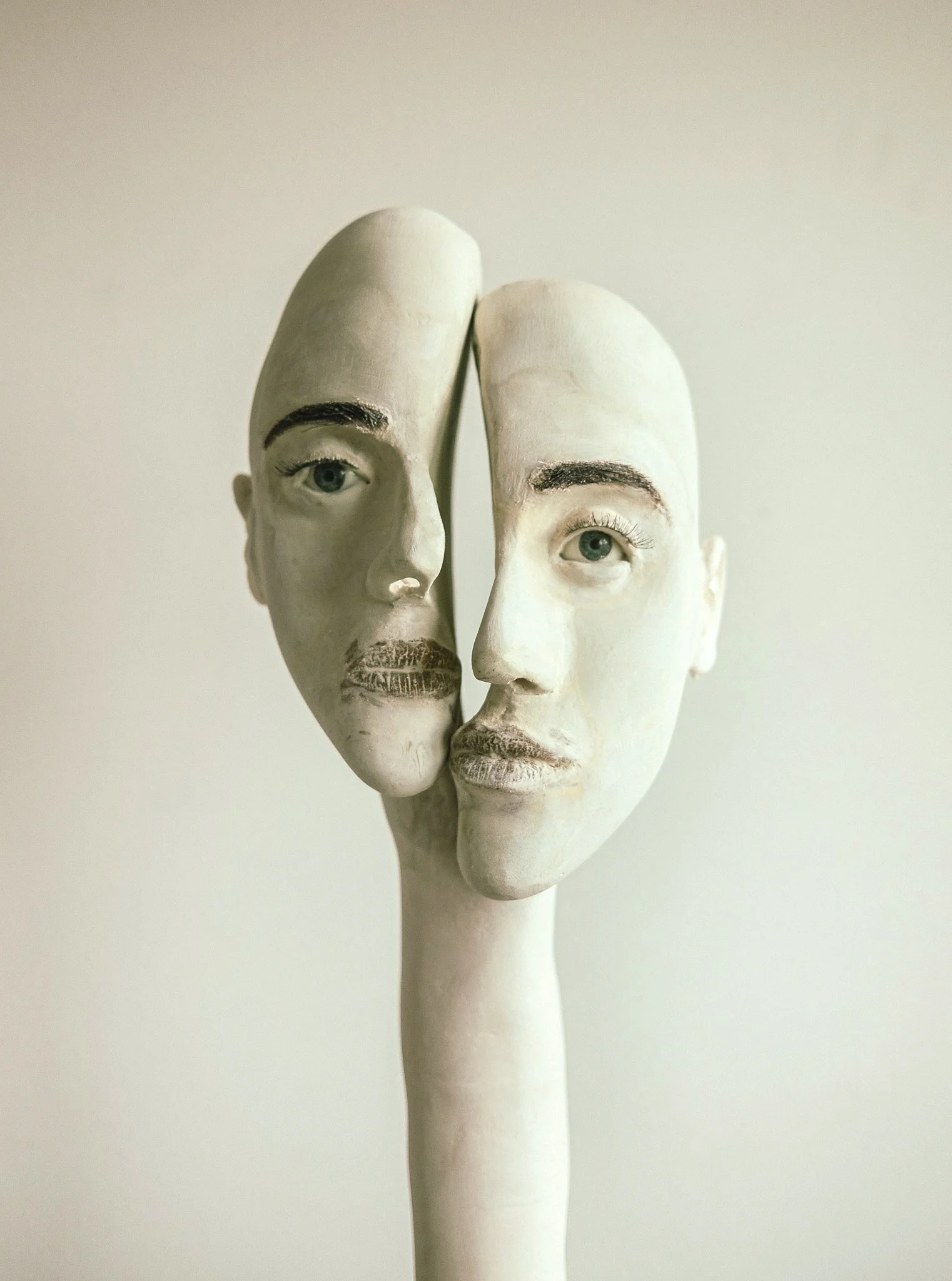 A sculpture of a face with two halves, one with a human face and the other with a mask, mounted on a tall, slender base.