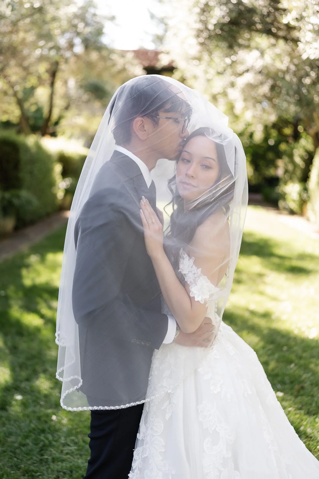A bride and groom in wedding attire embracing outdoors under a sheer veil, with greenery and trees in the background on a sunny day.
