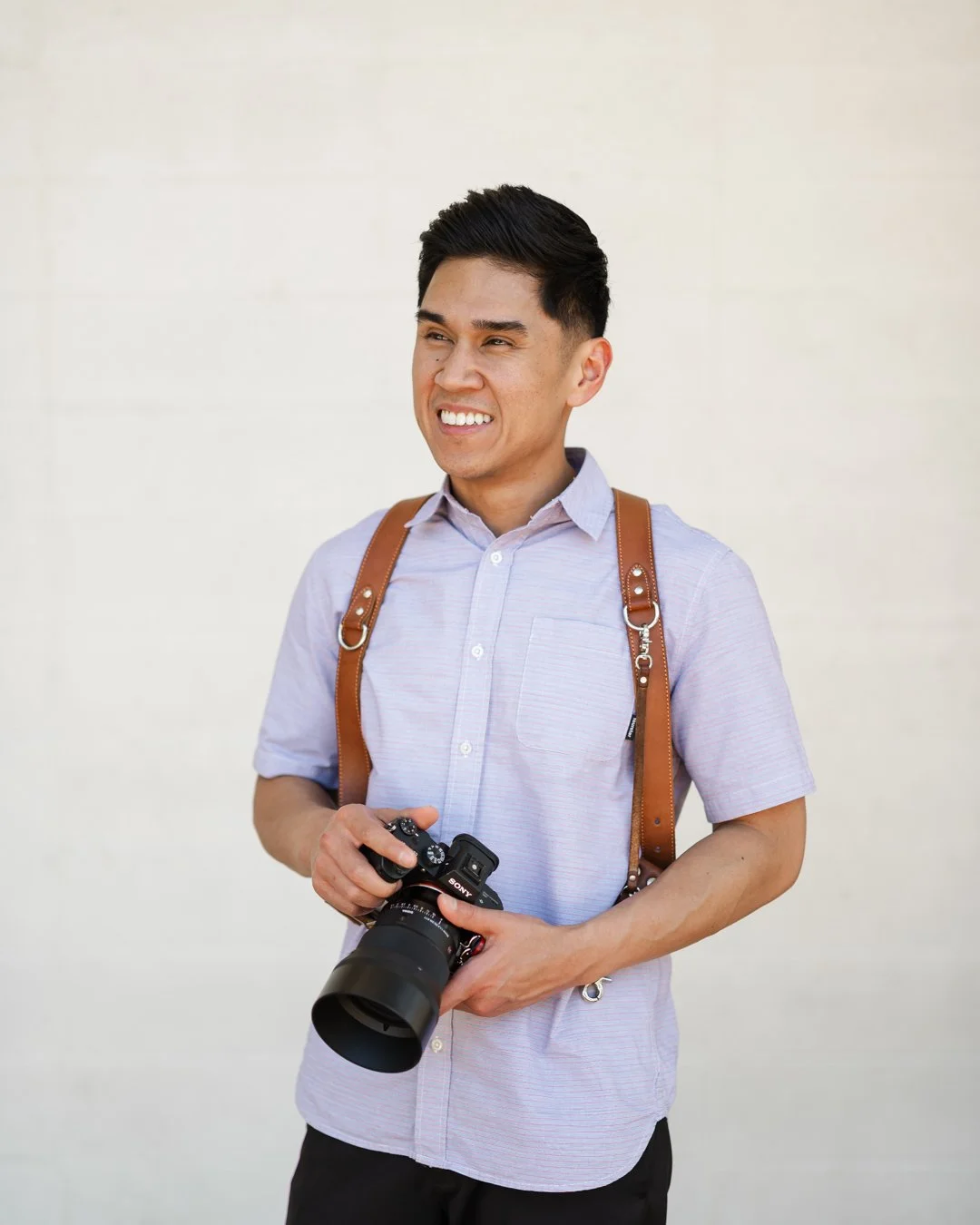 A young man with black hair, wearing a light purple button-up shirt, holding a DSLR camera and smiling, standing against a plain light-colored background.