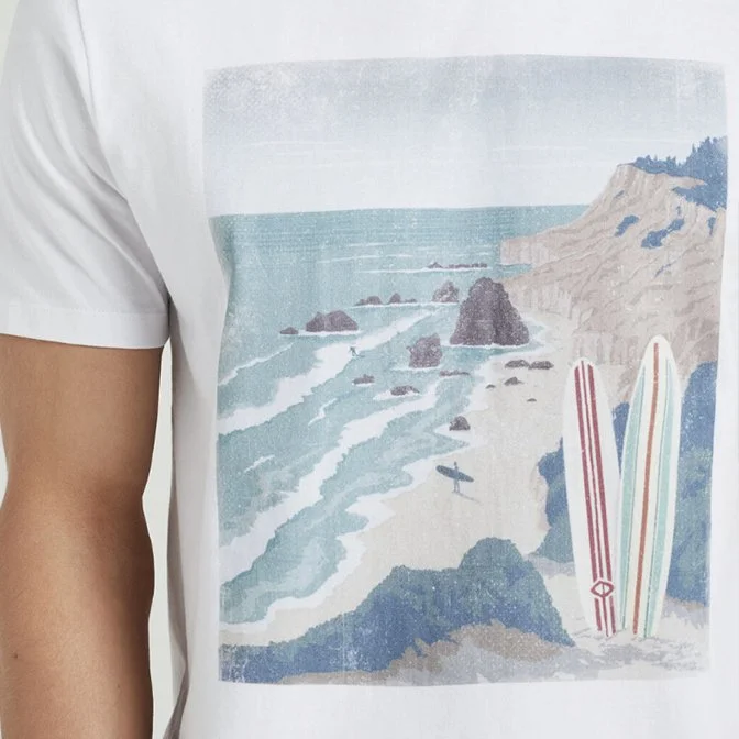 Close-up of a white t-shirt with a printed scene of a beach with waves, rocks, cliffs, and two surfboards resting at the shore.