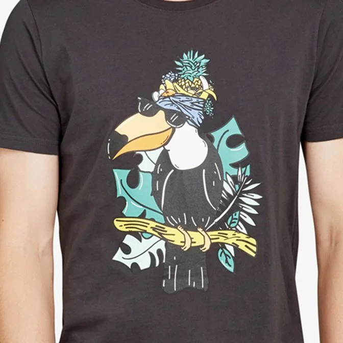 A person wearing a black T-shirt with a graphic of a toucan wearing sunglasses, a bandana, and a pineapple on its head, sitting on a branch surrounded by tropical leaves.