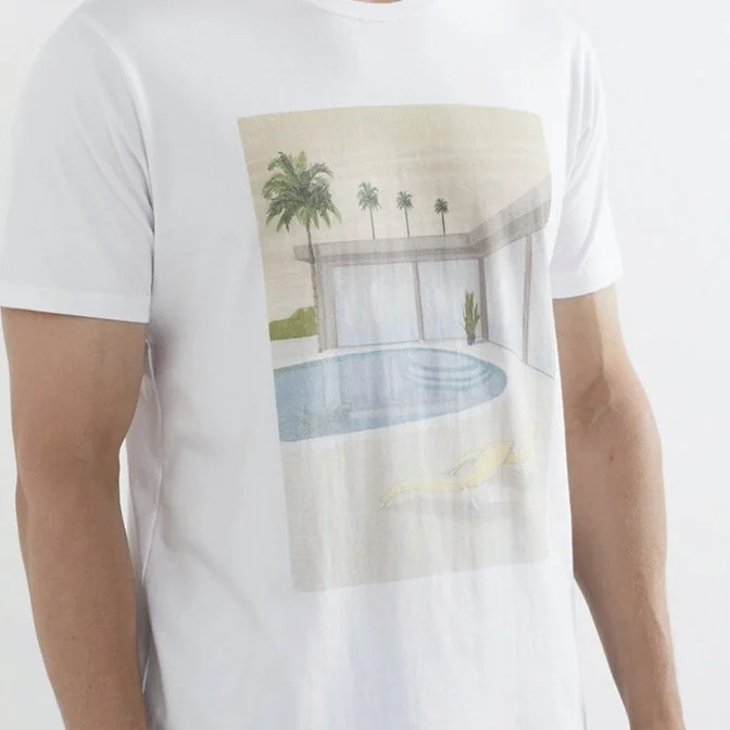 White T-shirt with a printed scene of a modern house with glass walls, a swimming pool, a yellow lounge chair, and palm trees in the background.