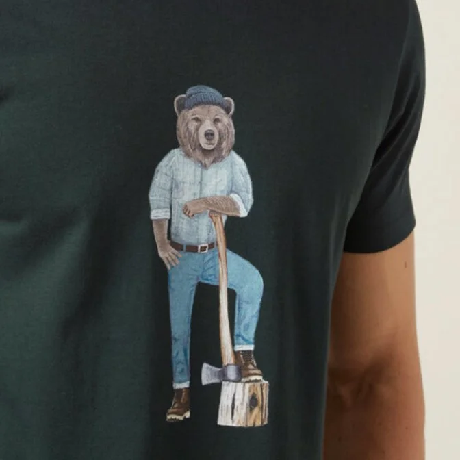 A person wearing a black T-shirt with a bear character illustration on it, featuring a bear with a human body, dressed in jeans and a shirt, holding an axe resting on a wooden stump.