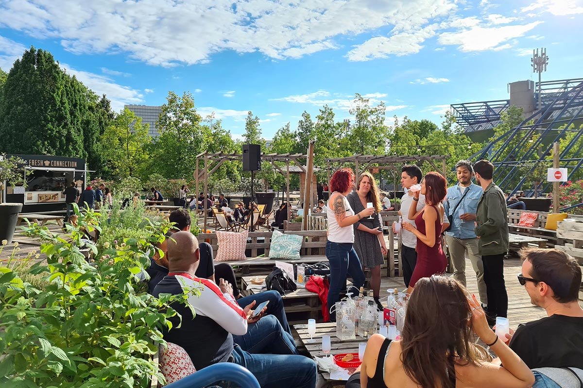 Personnes socialisant dans un espace extérieur, avec arbres et ciel bleu, buvant et discutant.