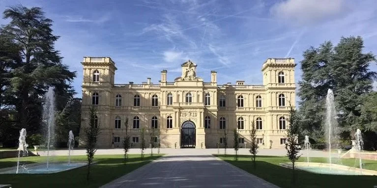 Photo d'un château néoclassique avec une façade symétrique, des fontaines devant et des arbres autour.