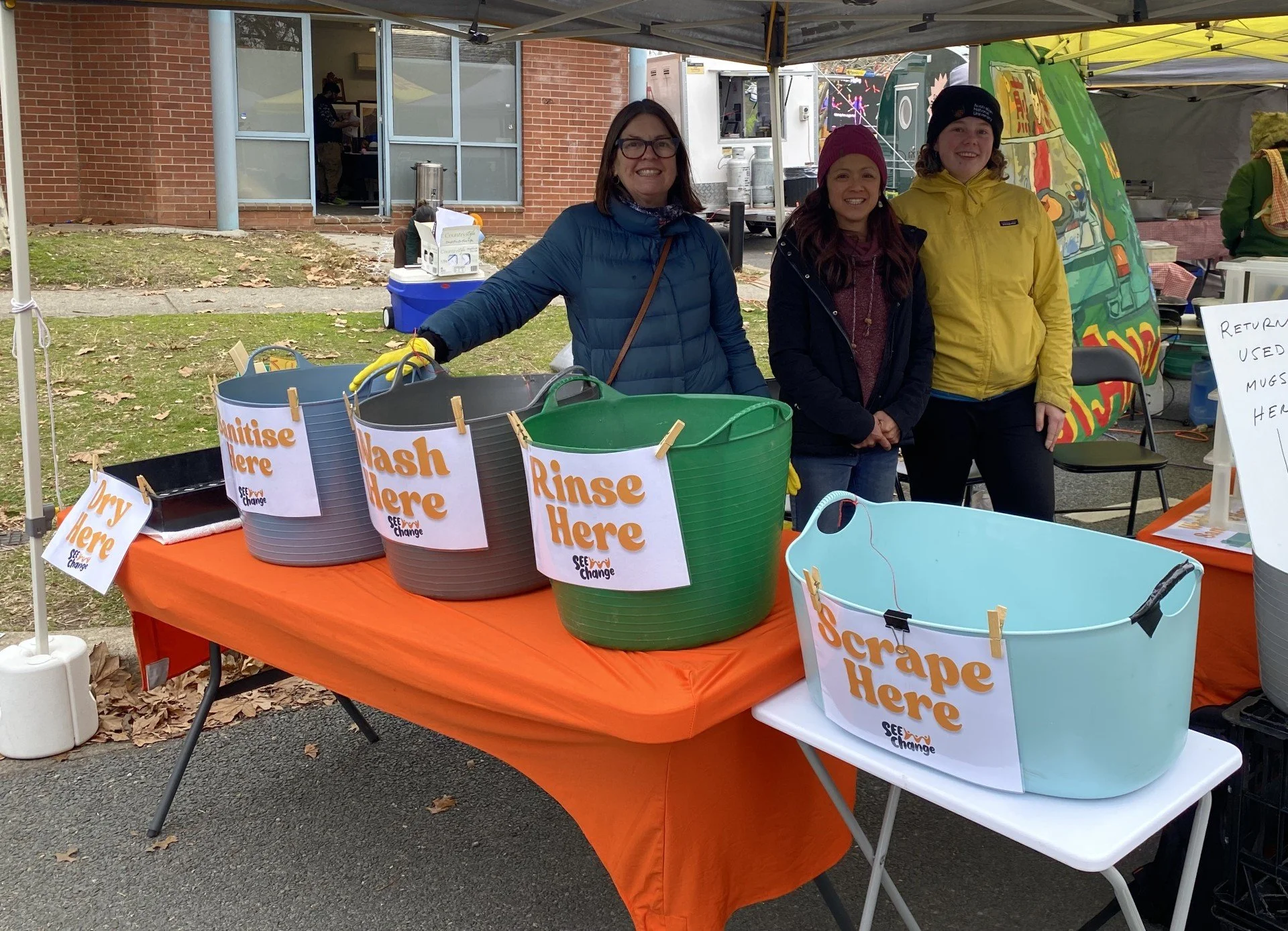 Three smiling women standing behind a line of four washing tubs