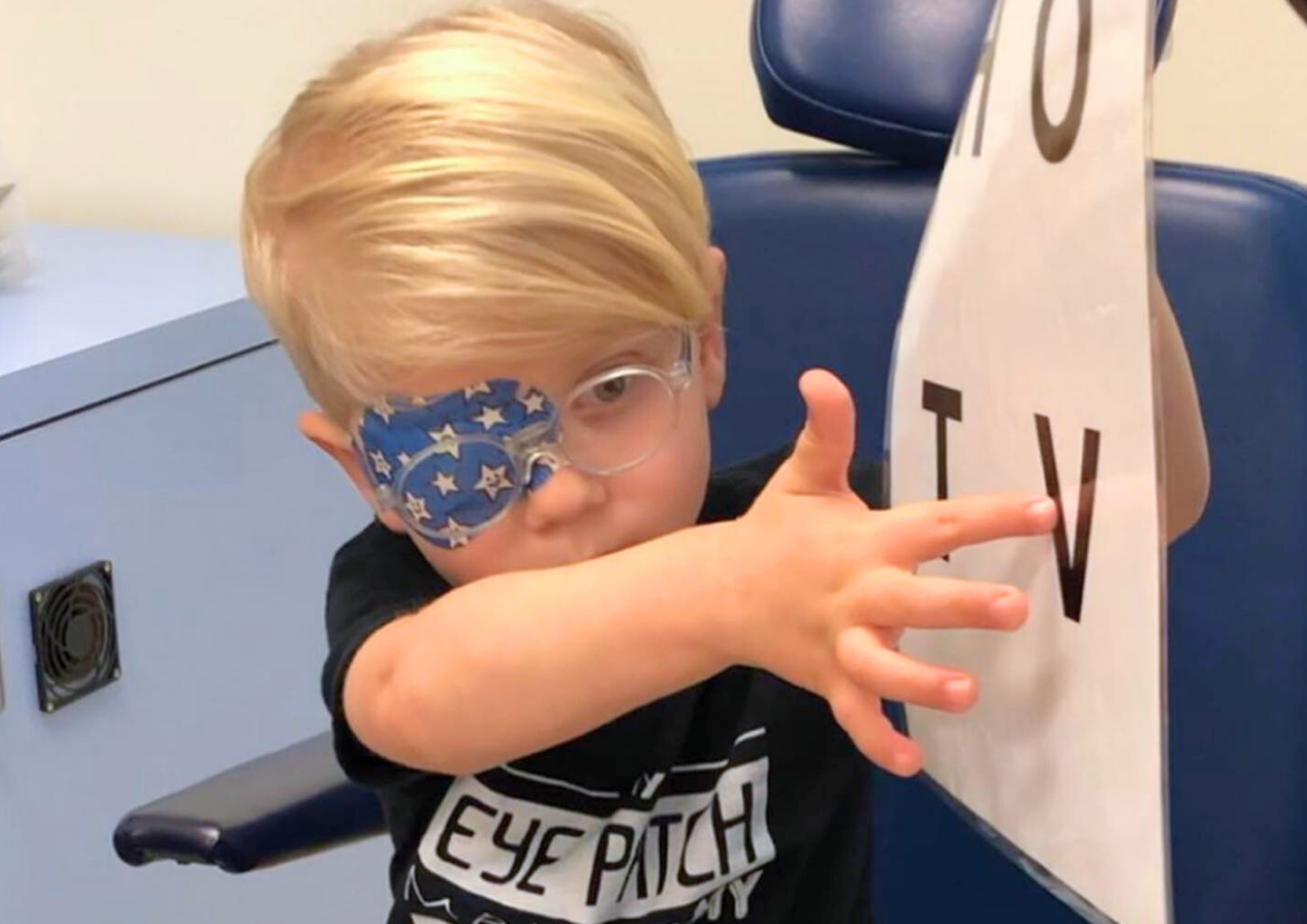 Preschool boy wears glasses, and has a patch on one eye. He is holding an eye test chart and pointing to a letter.