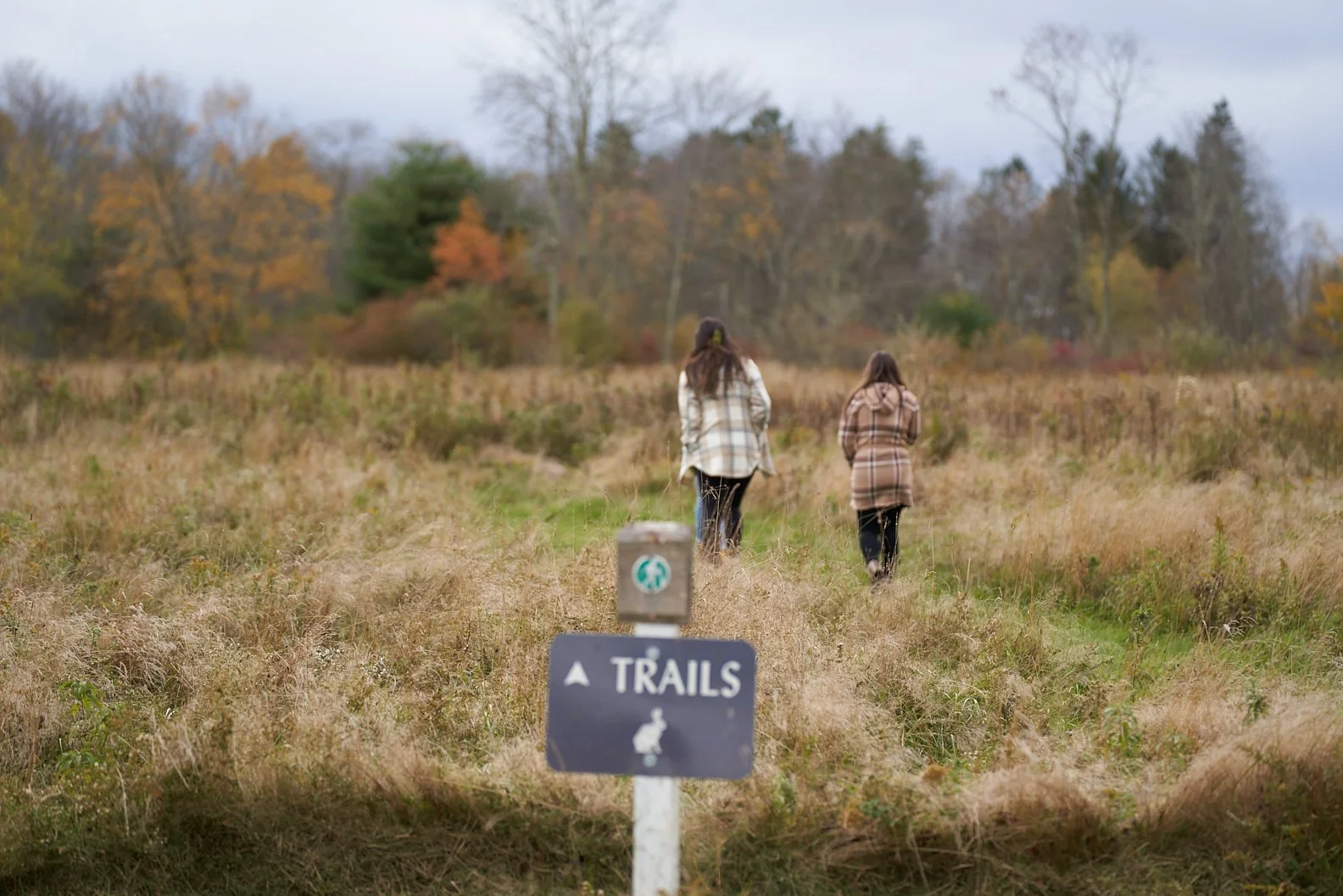 Two women are walking along a trail in a grassy field during the fall. They are dressed in plaid coats, and the sky is overcast. A sign in the foreground indicates this is a trail area.