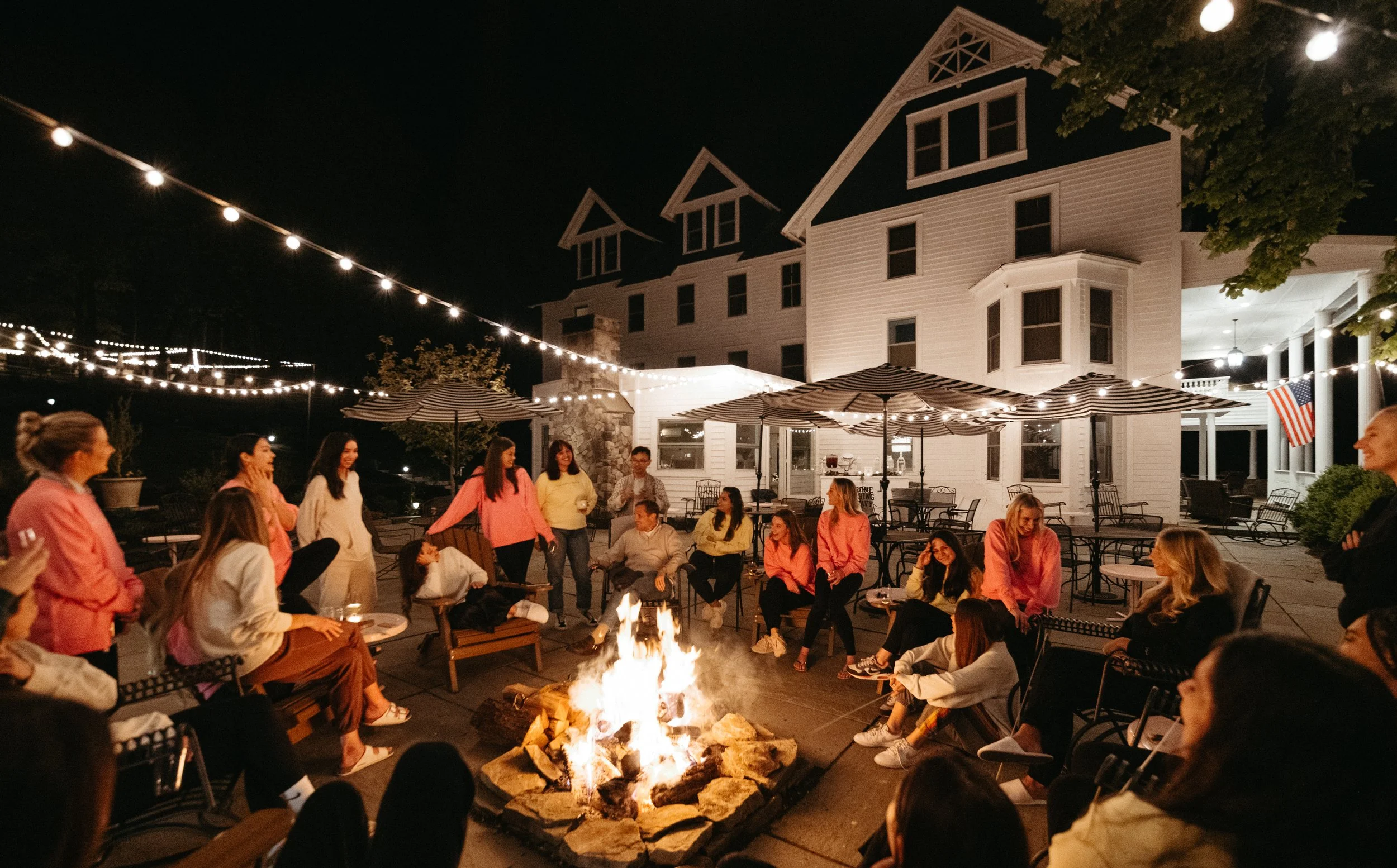 Nighttime outdoor gathering around a fire pit at a house with string lights, patio furniture, and a group of people socializing.