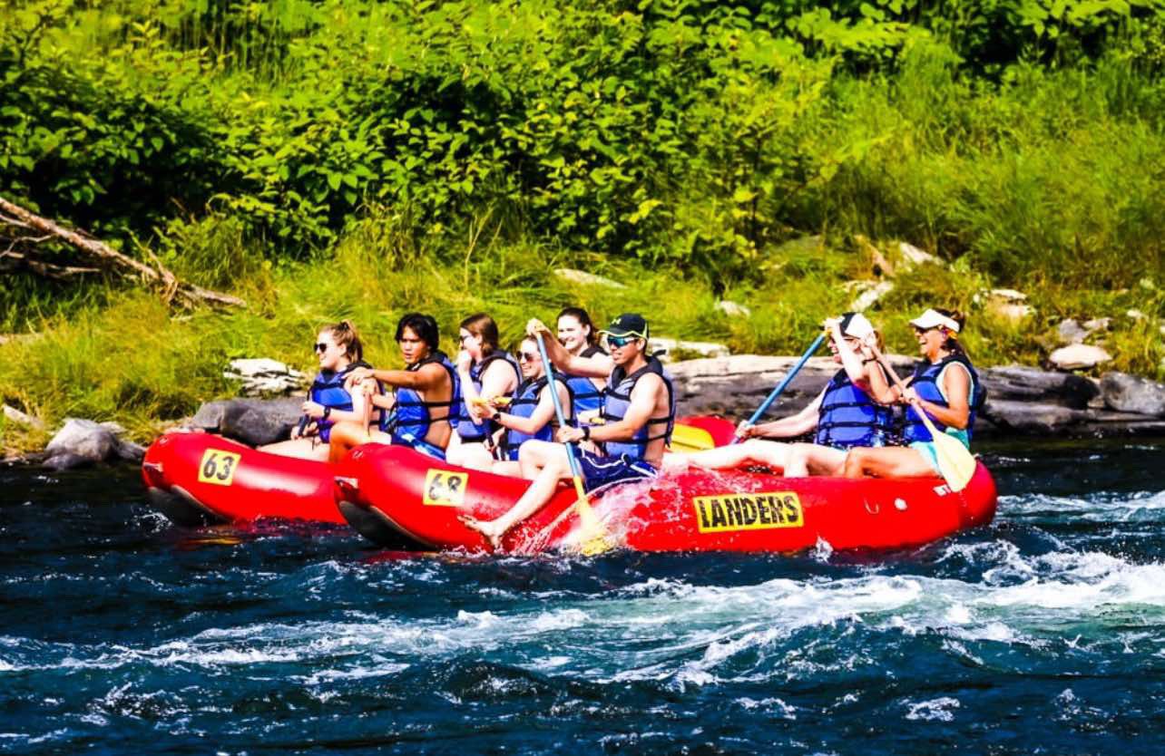 A group of people in a red inflatable raft paddling on a river surrounded by green trees and rocks, enjoying a summer outdoor activity.